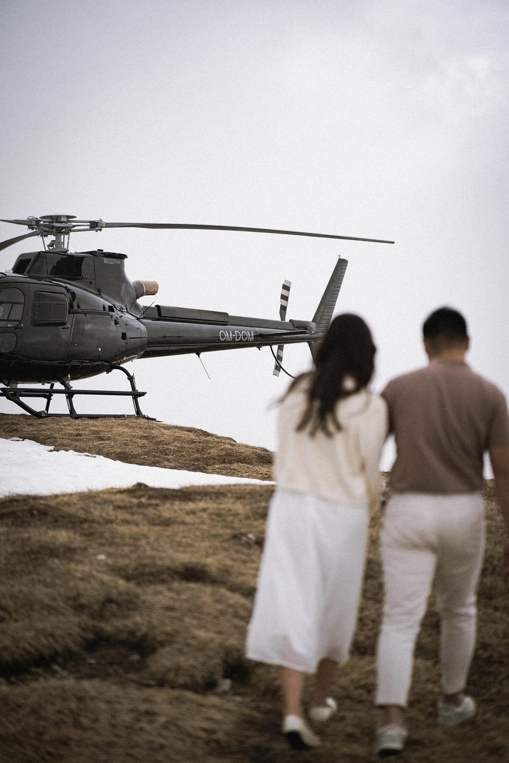 Ein schwarzer Hubschrauber auf einem Hügel mit zwei Menschen im Vordergrund, die sich an den Händen halten und den Hubschrauber betrachten, bei bewölktem Himmel.