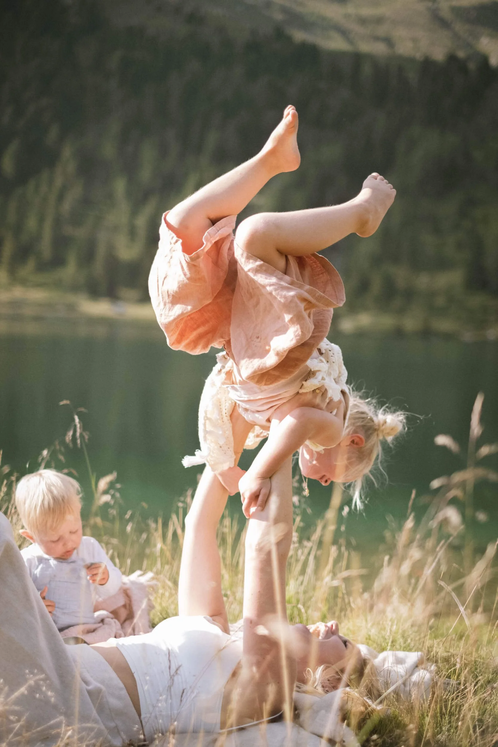 Eine Frau liegt auf dem Rücken im Gras, hebt ein Mädchen hoch und es befindet sich im Handstand auf ihrer Hand, während ein kleiner Junge im Hintergrund im Gras sitzt.