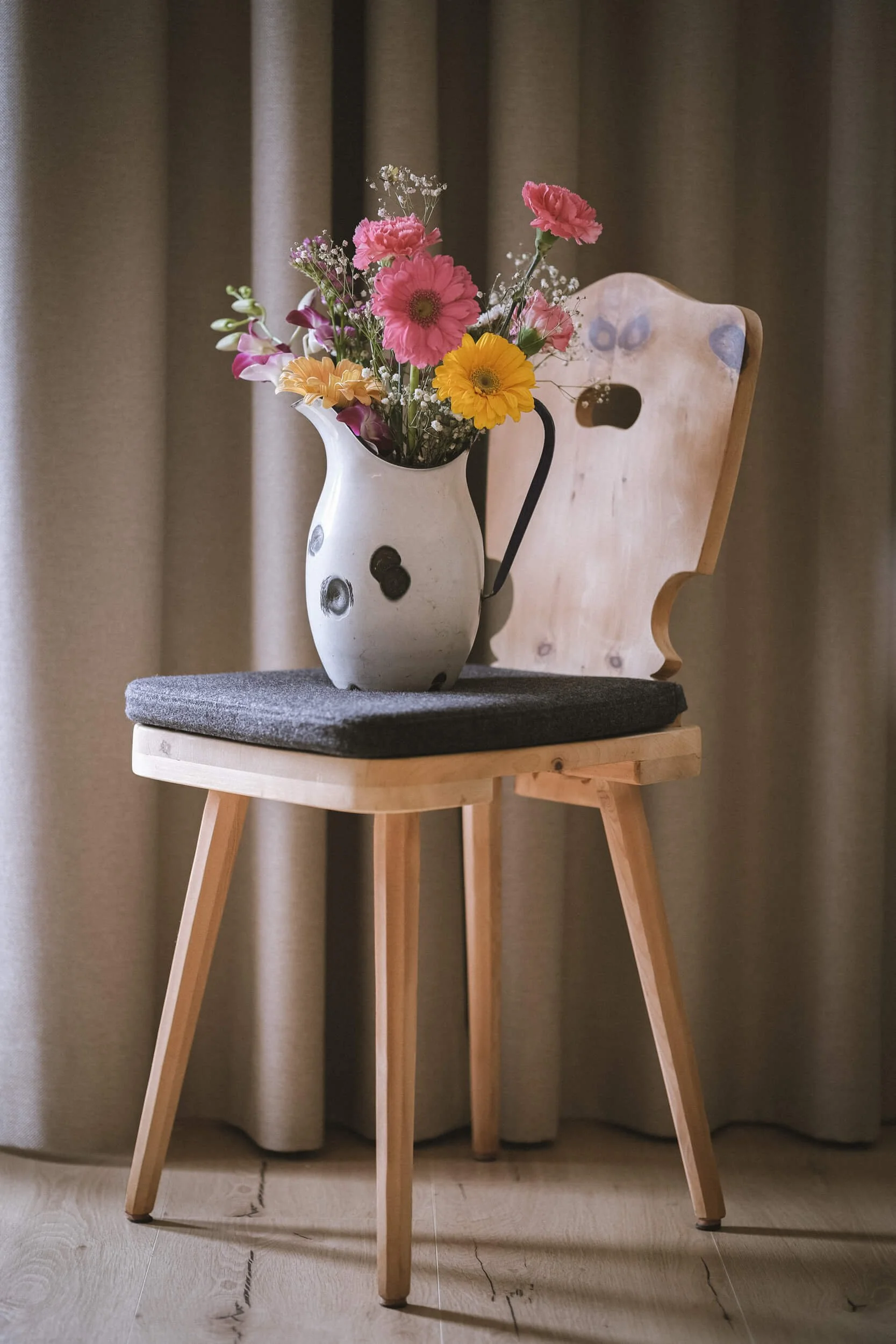 Ein Blumenstrauß in einer weißen Vase mit schwarzen Flecken steht auf einem Stuhl. Der Stuhl hat einen Holzrahmen und eine graue Sitzfläche.