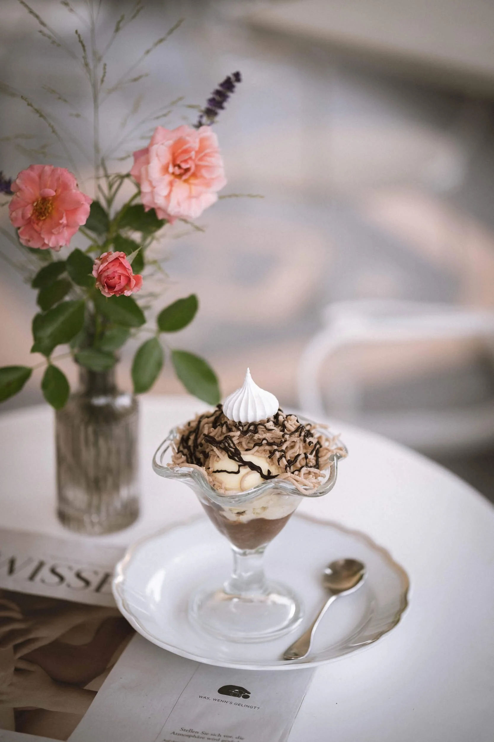 Ein Eisbecher mit Schlagsahne, Schokoladenraspeln und Schokoladensauce auf einem weißen Teller, daneben eine Silberlöffel, im Hintergrund eine Vase mit rosa und lila Blumen auf einem Tisch.