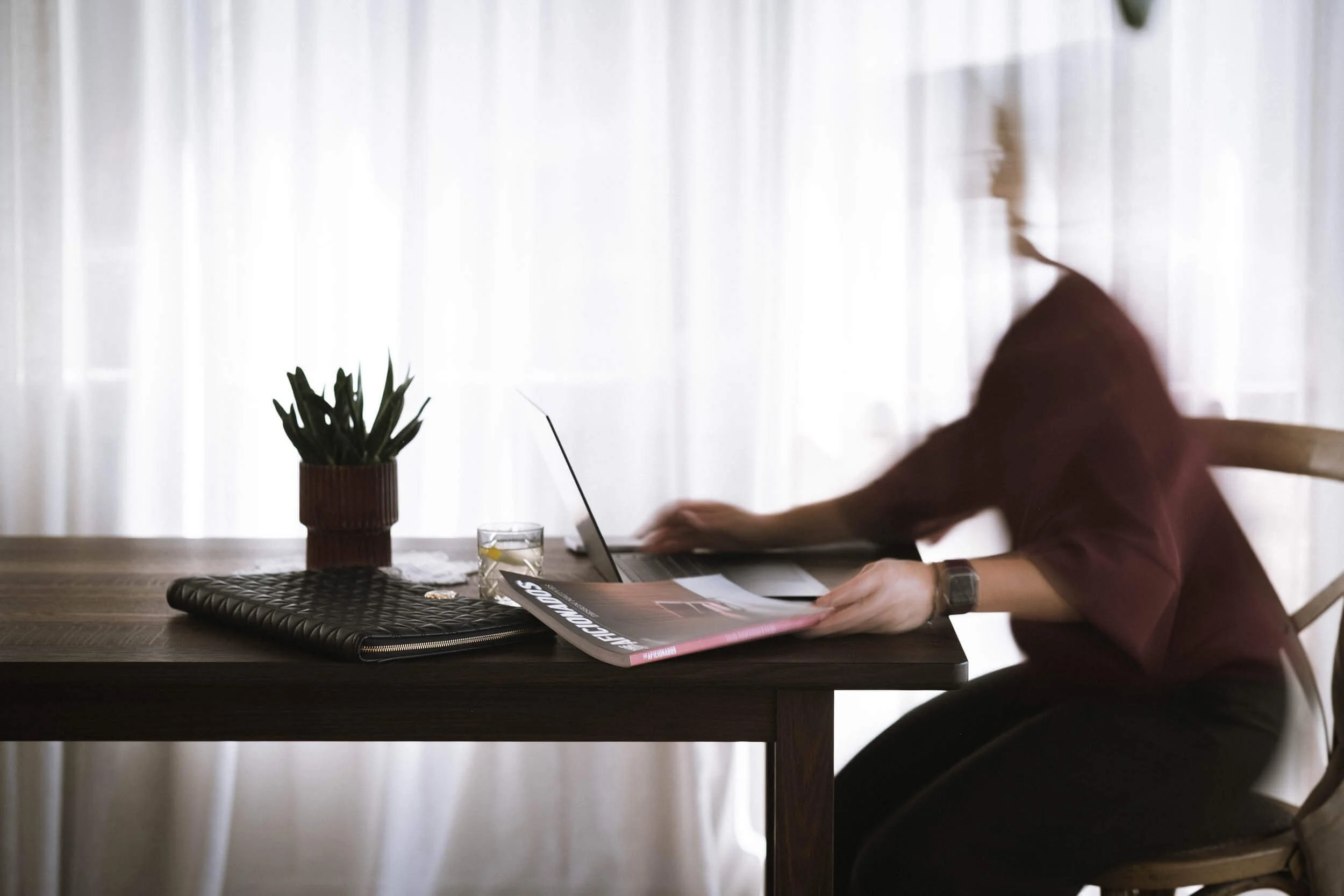 Person sitzt an einem Tisch, arbeitet an einem Laptop und liest eine Zeitschrift, mit einer Tasse Wasser und einer Pflanzen im Hintergrund.