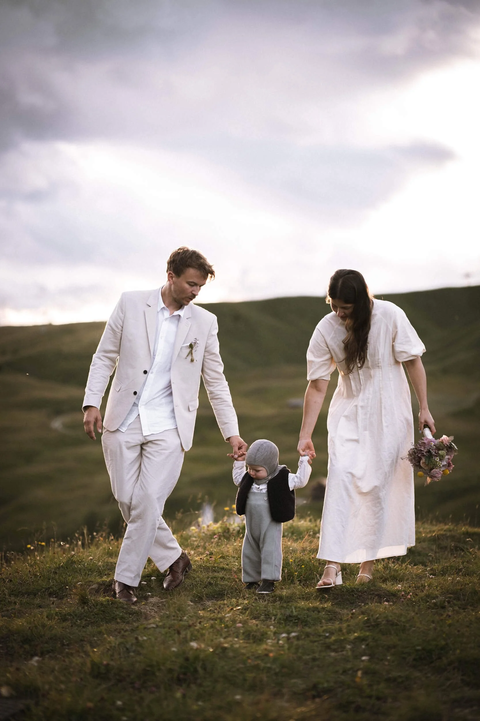 Ein Mann und eine Frau spazieren mit einem kleinen Kind auf einer grünen Wiese im Freien bei Sonnenuntergang, das Kind hält die Hände der Erwachsenen, die abseits von Gebäuden und Straßen sind.