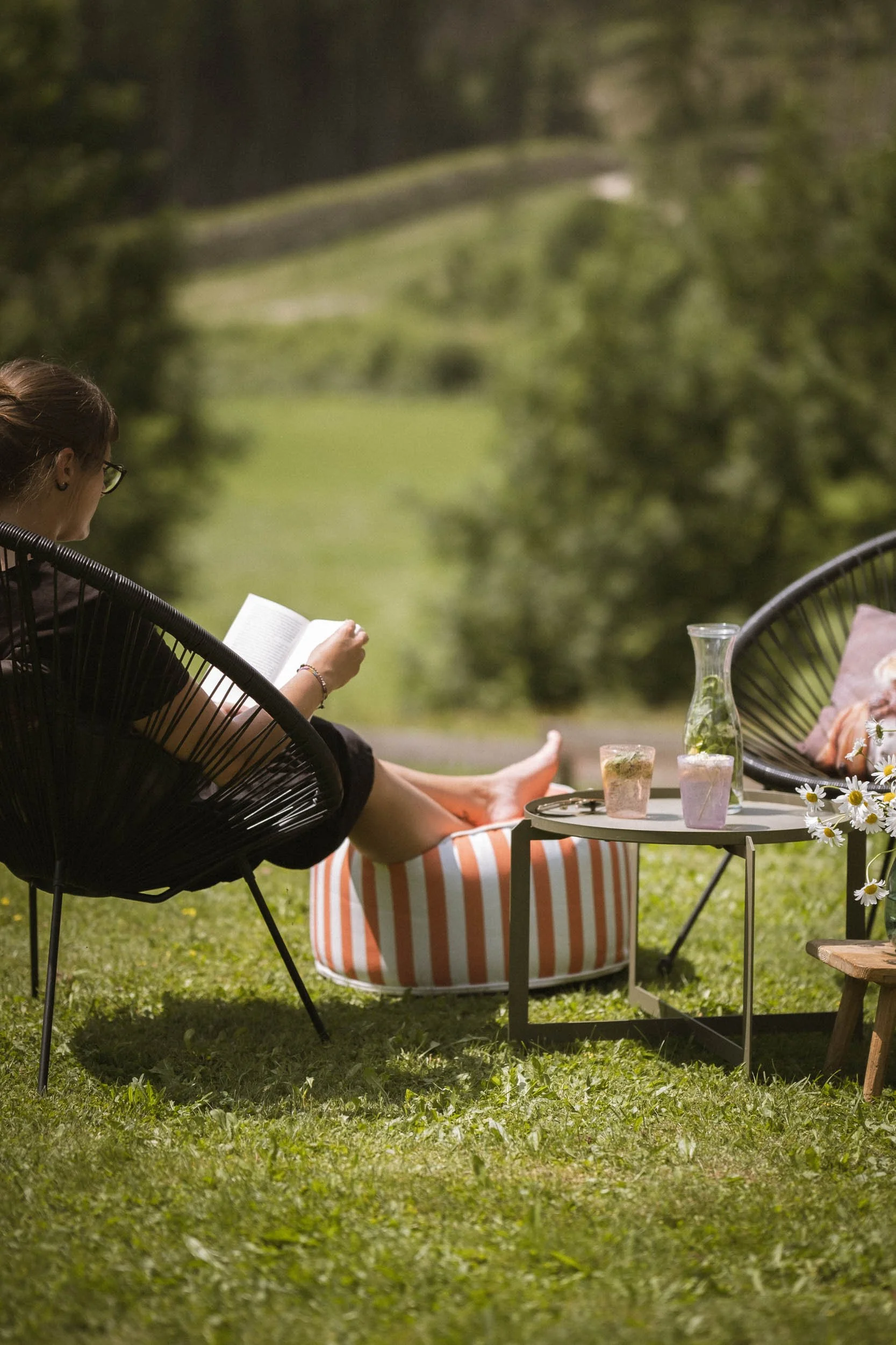 Eine Frau liest ein Buch im Garten, während sie auf einem gestreiften Sitzsack sitzt. Auf einem kleinen Tisch stehen Getränke, und im Hintergrund ist eine grüne Landschaft zu sehen.