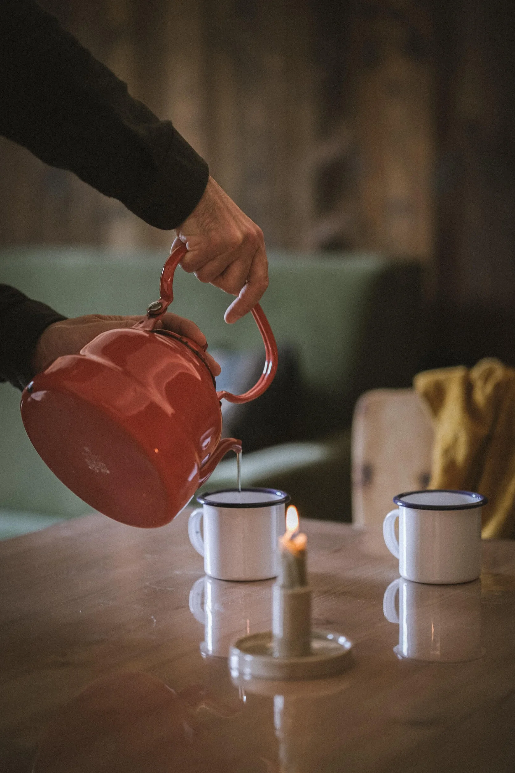 Eine Person gießt Wasser aus einer roten Kanne in eine weiße Tasse auf einem Holztisch, mit einer brennenden Kerze zwischen zwei Tassen.