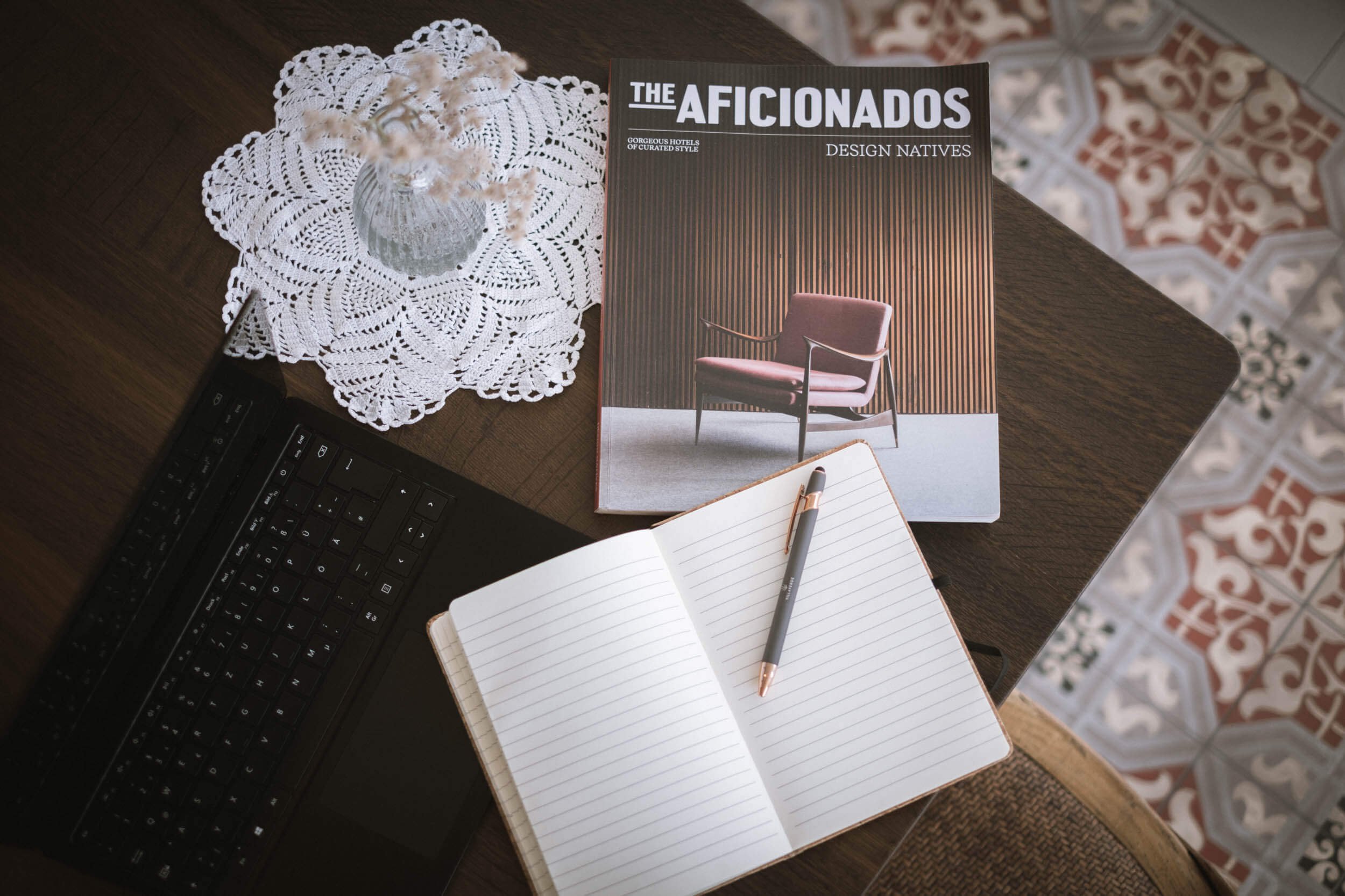 Ein Arbeitsplatz mit einer offenen linierten Notizbuch, einem schwarzen Kugelschreiber, einem Laptop, einer weißen gehäkelten Tischdecke mit Vase, einem Magazin mit dem Titel "The Aficionados" und einem roten Stuhl im Hintergrund, auf einem Holztisch