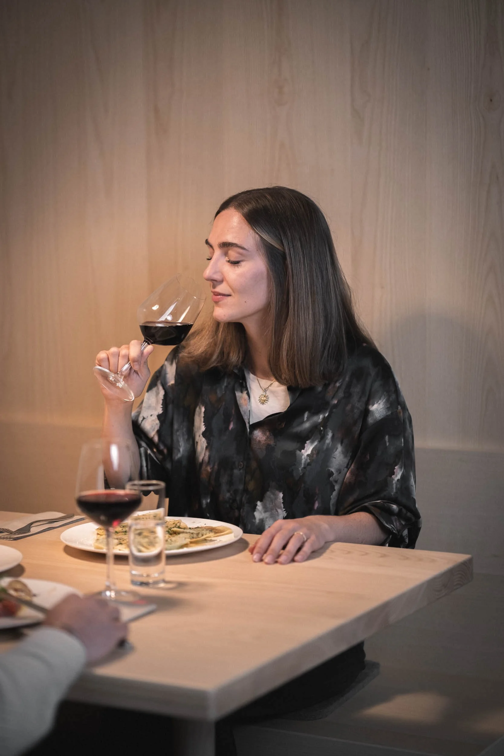 Frau mit braunen Haaren, die ein Glas Rotwein hält, sitzt am Tisch mit Essen, auf einem hellen Holztisch, vor einem Holzpaneelwand