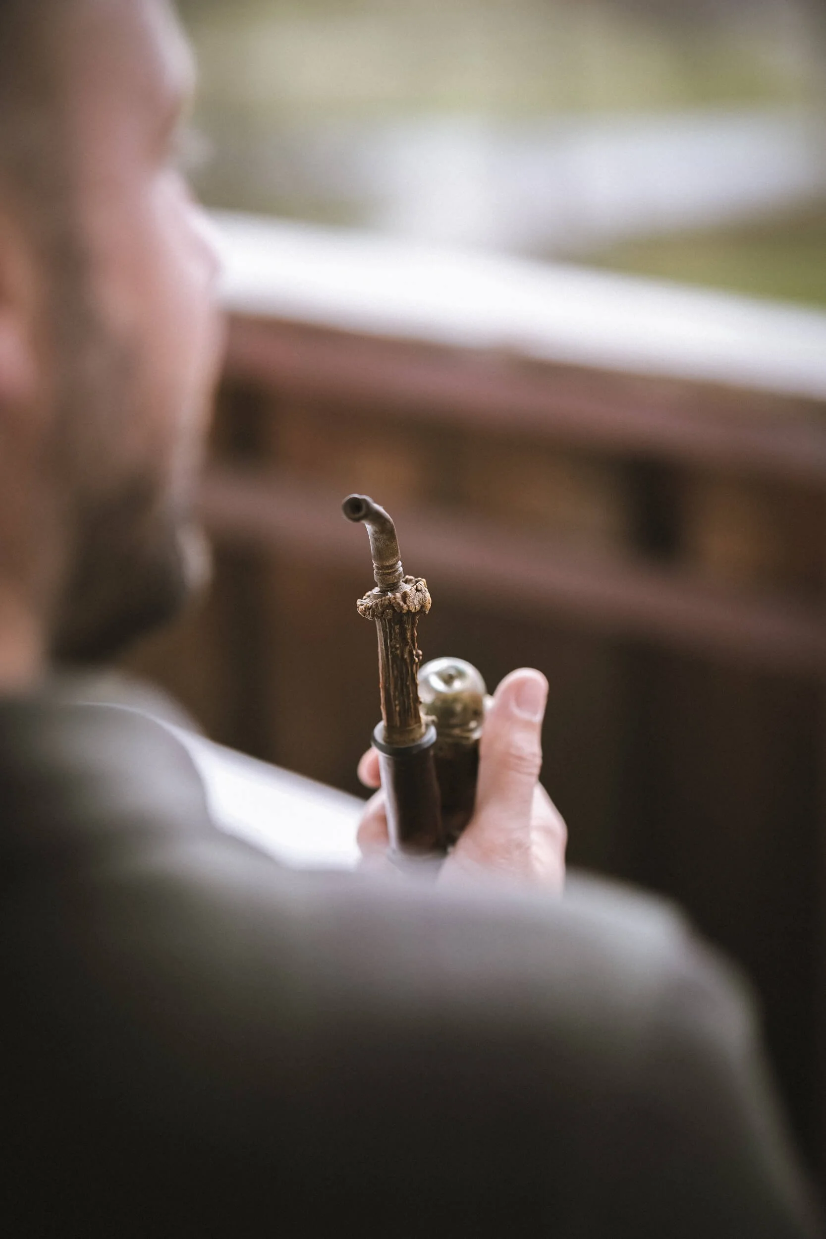 Ein junger Mann mit Bart hält eine antike Pfeife und eine Tabakdose in der Hand, sitzend in einem Raum mit Holzsitzbank.