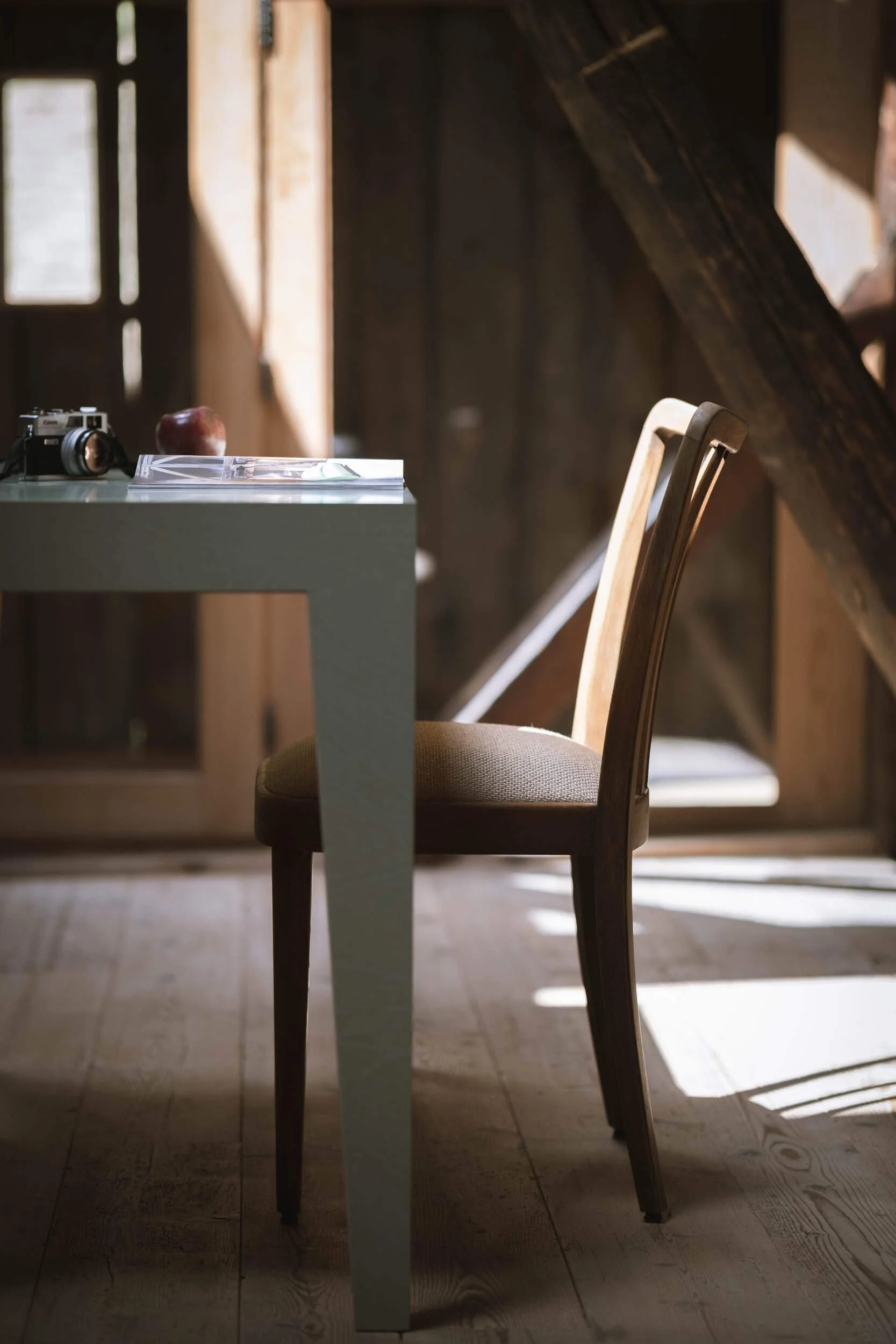 Ein Platz an einem Tisch mit einem Stuhl, einer Kamera, einem Apfel und Zeitschriften, in einem Raum mit Holzwänden und natürlichen Lichtstrahlen.