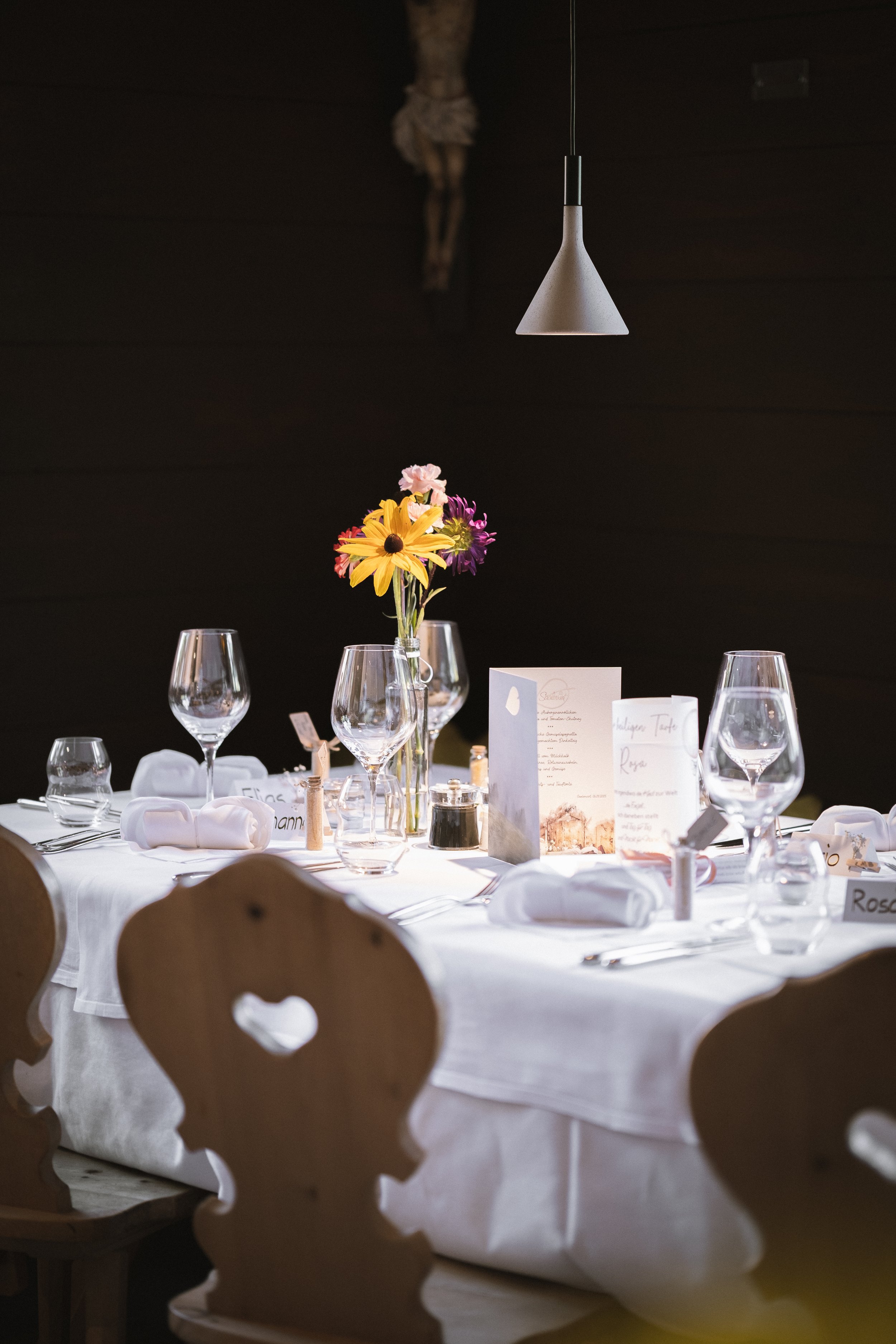 Ein festlich gedeckter Tisch mit weißen Tuch, mehreren Weingläsern, Besteck und einer kleine Vase mit bunten Blumen in einem dunklen Raum.