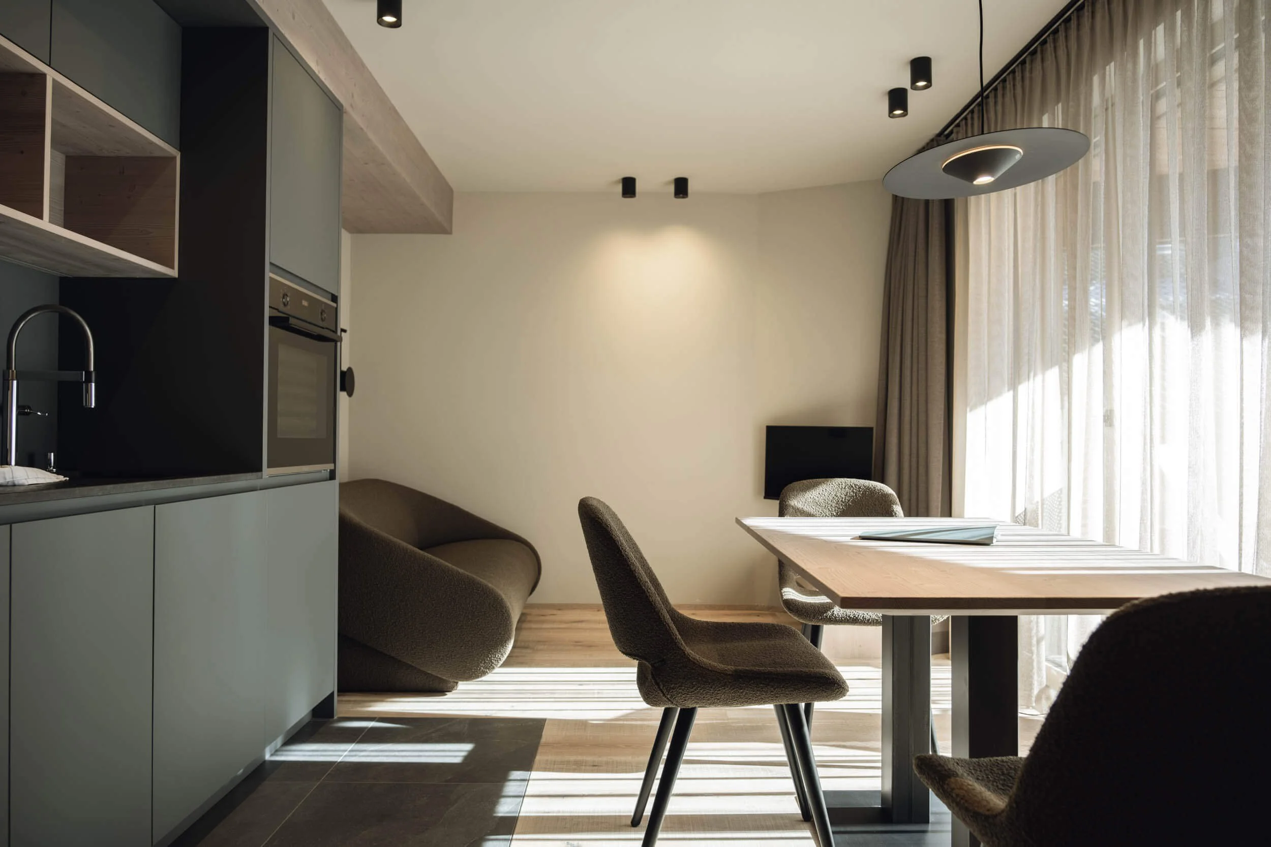 Moderne Wohnung mit Esstisch, Stühlen und Sofa vor großen Fenstern mit Gardinen, hellem Boden und minimalistischem Design.