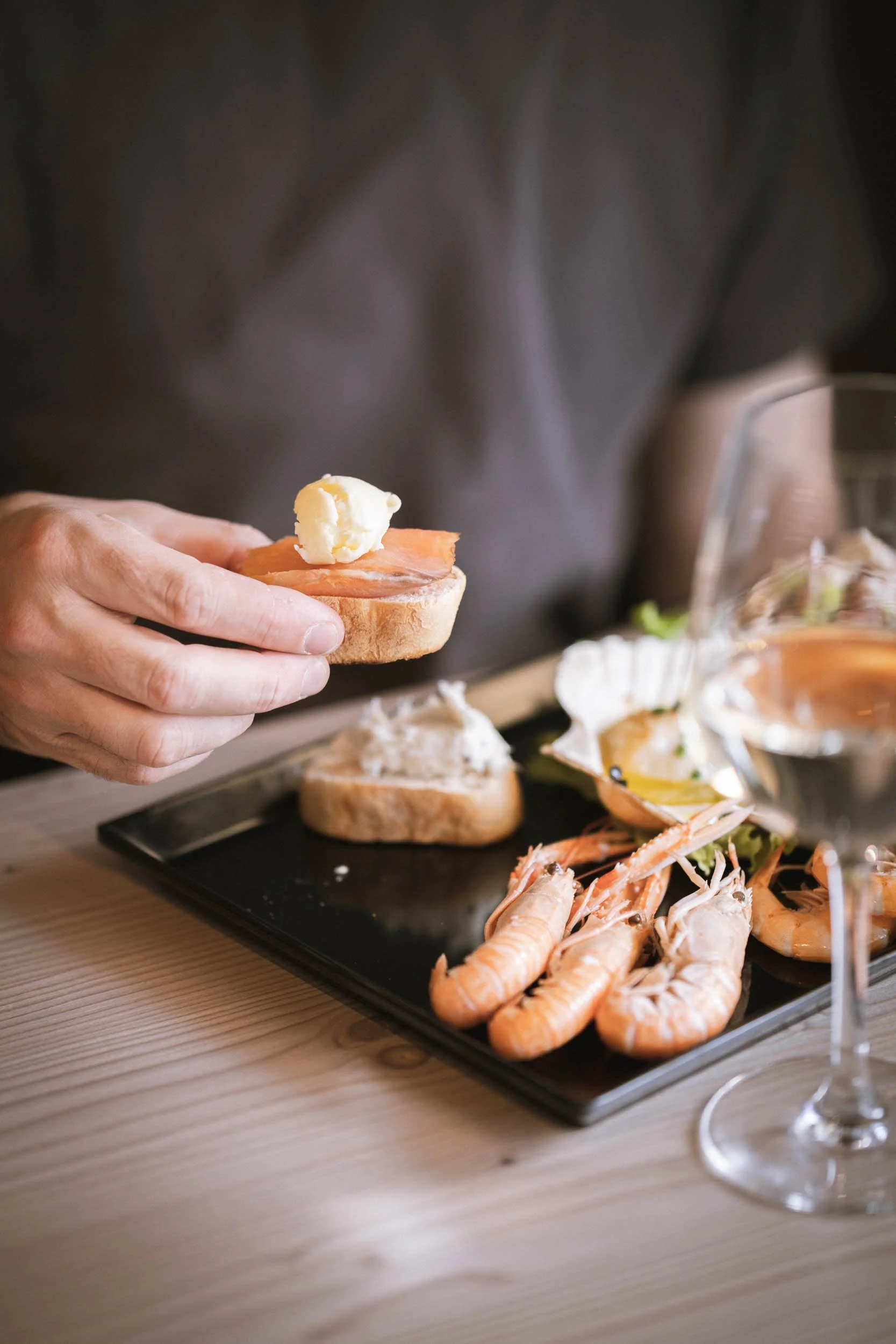 Nahaufnahme einer Hand, die eine belegte Brotscheibe mit Butter hält, neben einem schwarzen Teller mit Meeresfrüchten, darunter Garnelen, und einem Glas Weißwein auf einem Holztisch.