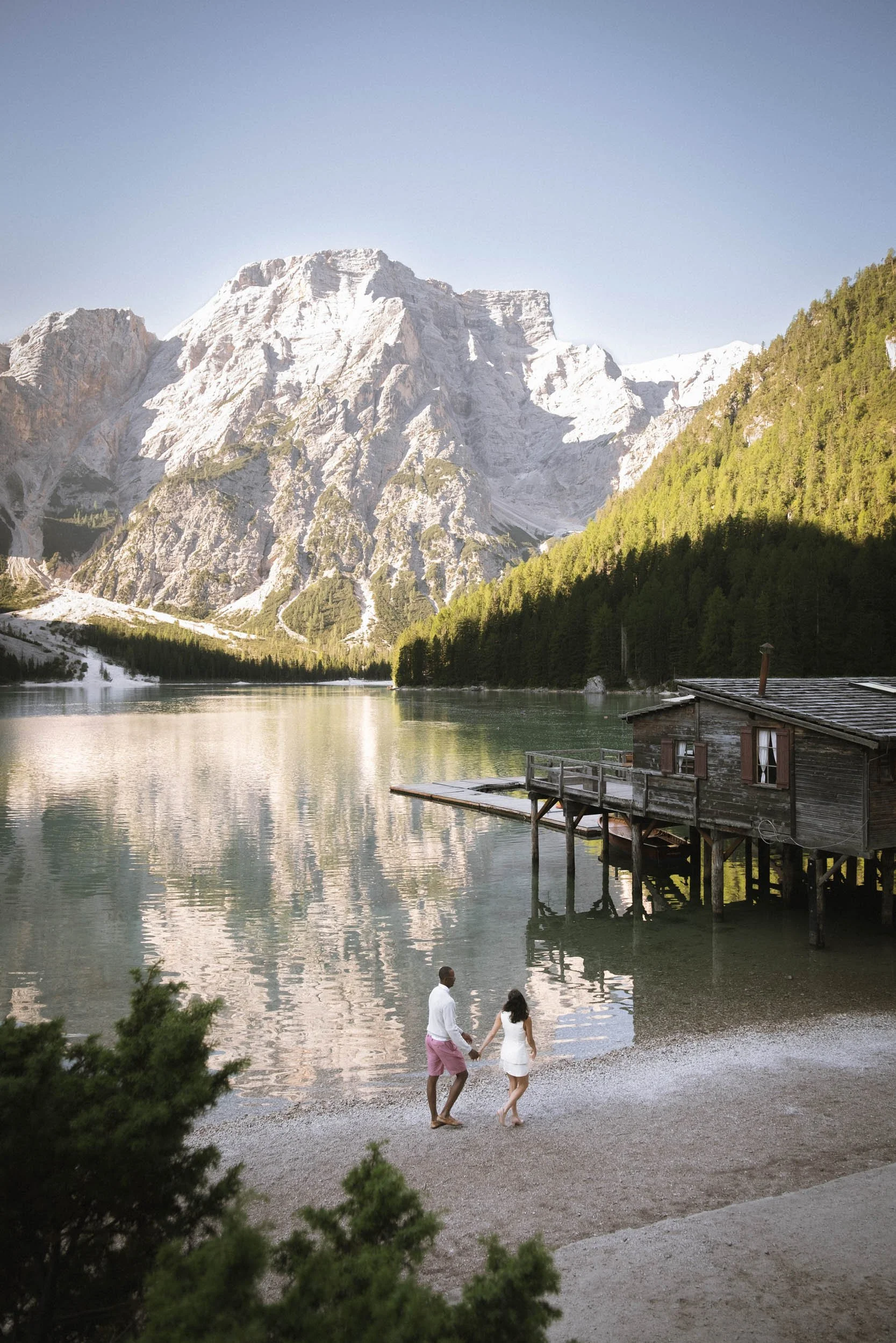 Ein Paar hält Händchen und geht am Ufer eines Sees im Gebirge spazieren, mit einer Holzhütte am Wasser und hohen Bergen im Hintergrund.