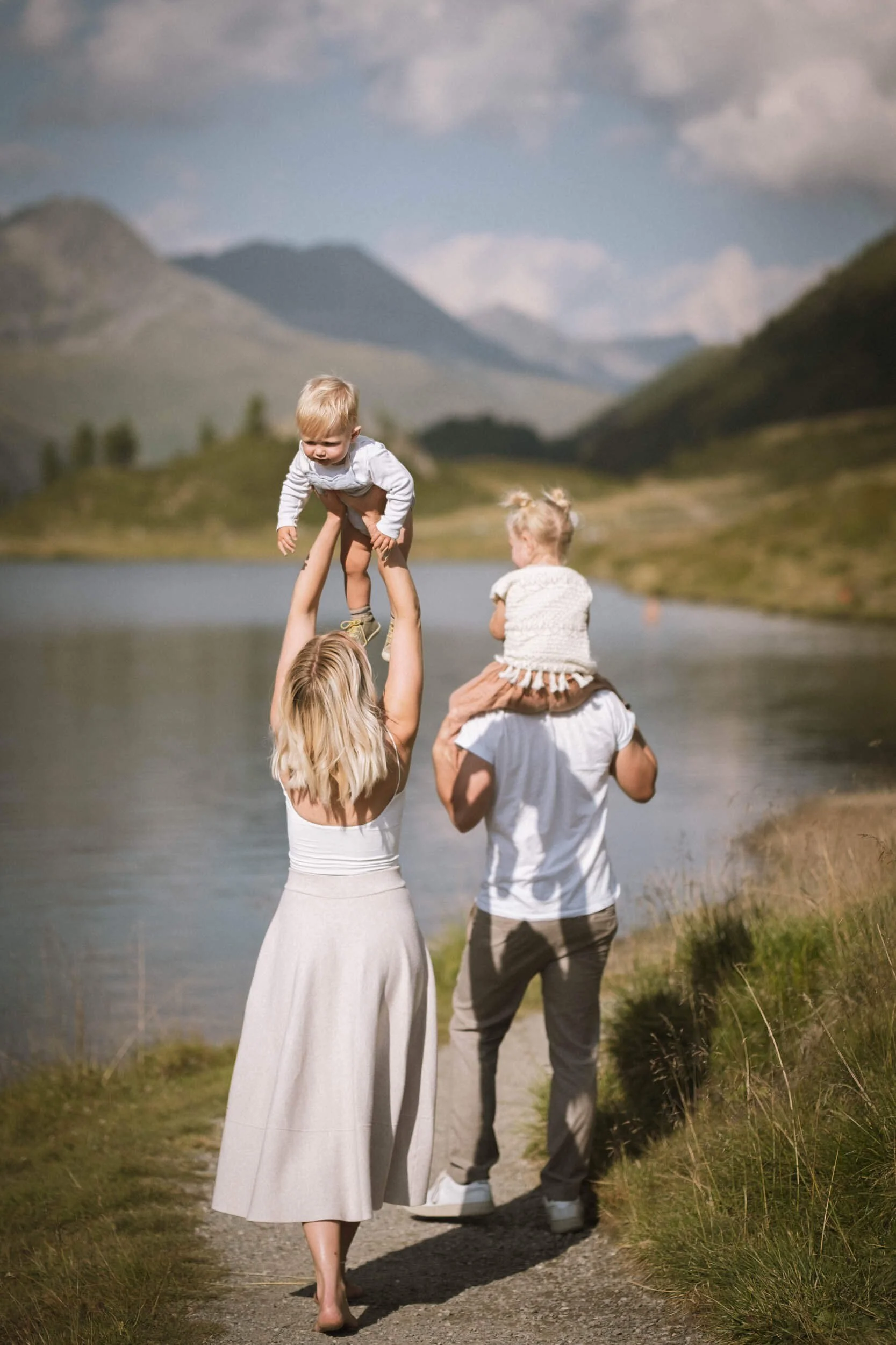 Eine Familie mit einer Frau, einem Mann und zwei kleinen Kindern, die am Ufer eines Sees in einer bergigen Landschaft spazieren gehen, wobei die Frau ein Kind hochhebt.