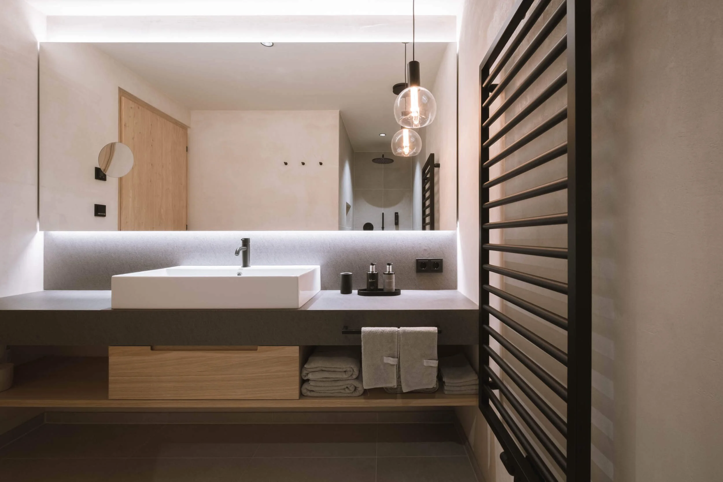 Moderner Badezimmer mit heller Wand, Spiegel, Waschbecken, schwarzen Armaturen, Handtüchern, beleuchteter Spiegel, schwarzer Heizkörper, Holzmöbel und Handwaschmitteln.