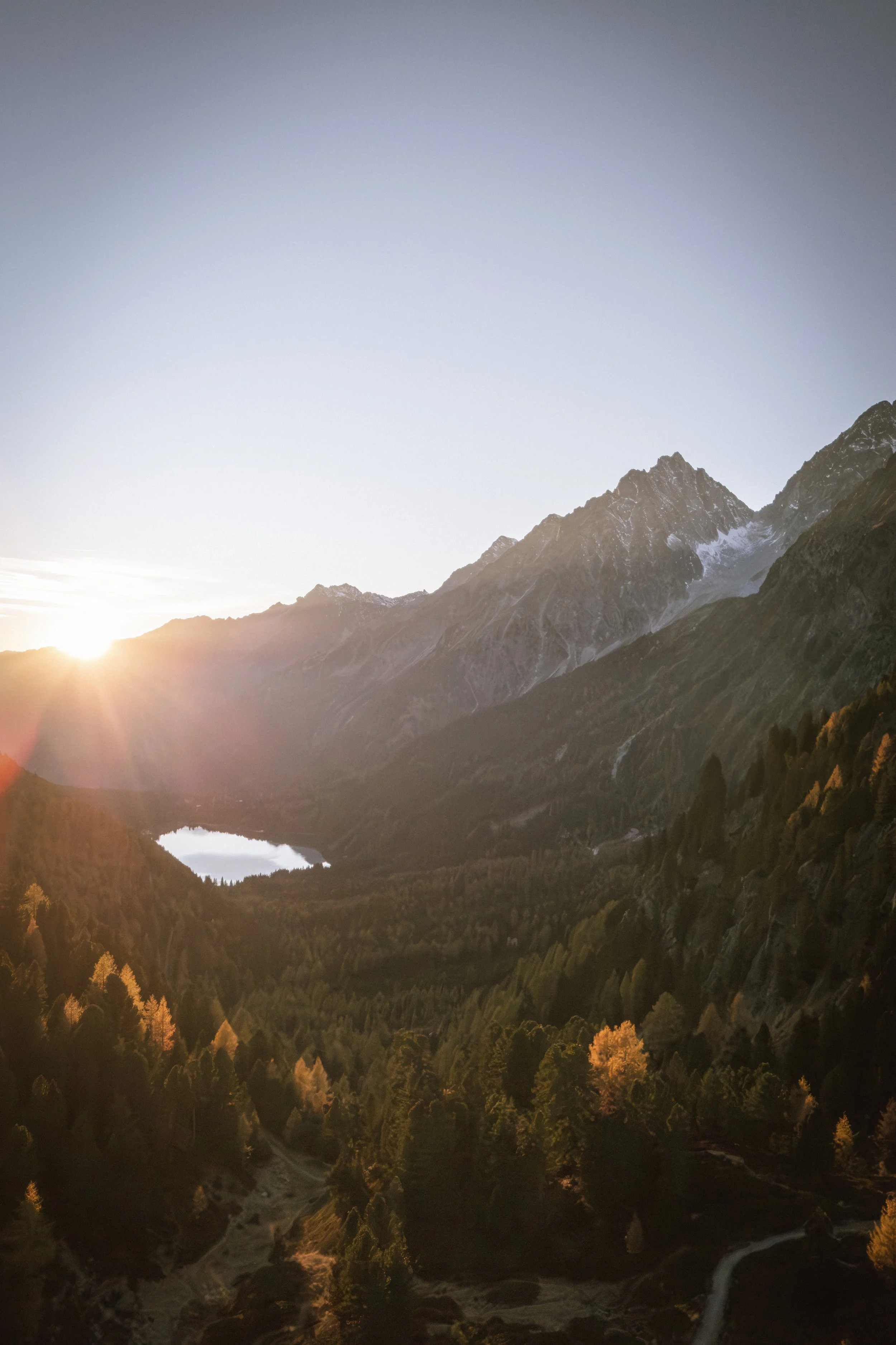 Berglandschaft mit einem See, bewaldeten Tälern und den verzweigten Wegen im Tal, Sonnenaufgang über den Bergen.