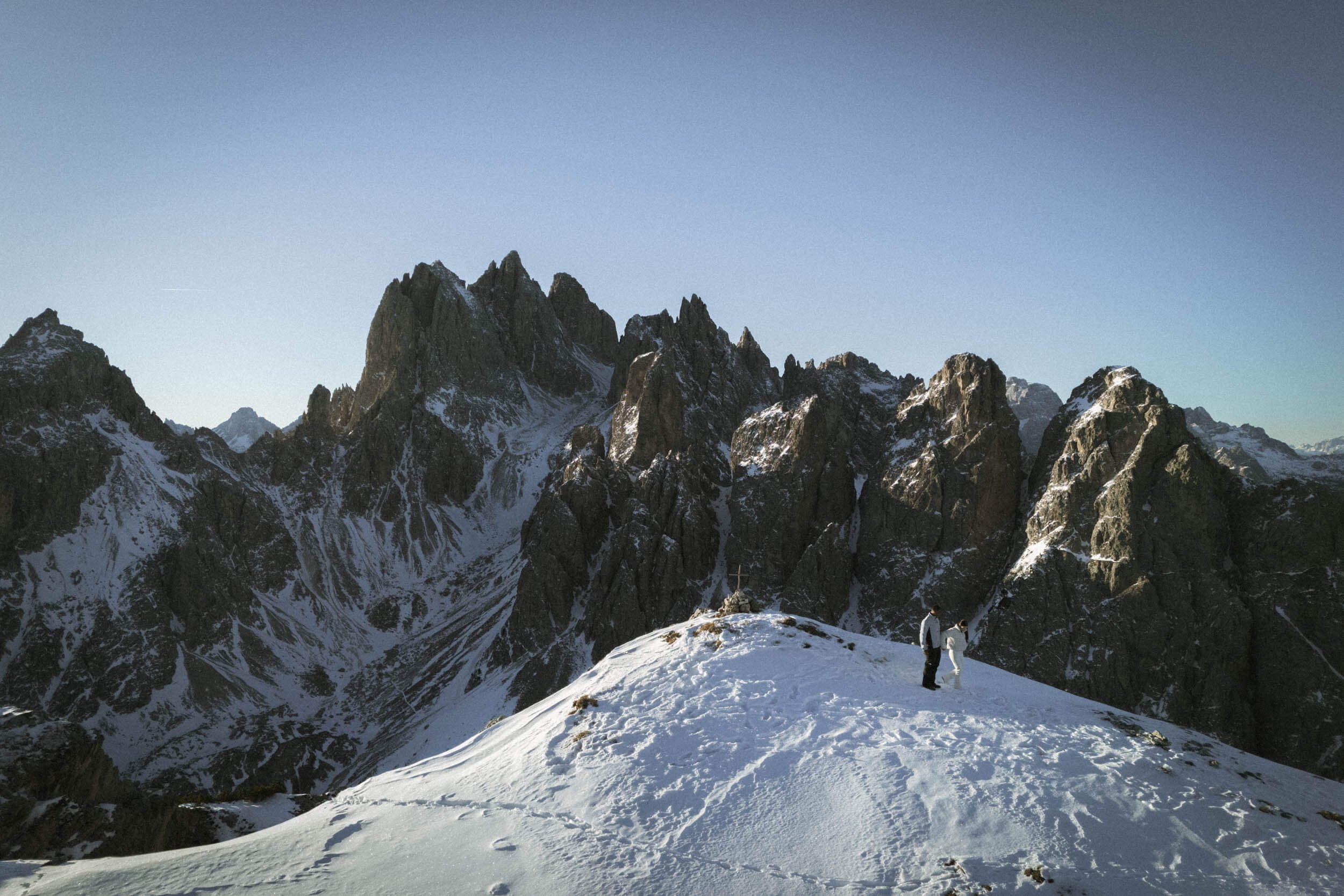 Zwei Menschen stehen auf einem schneebedeckten Berg mit Blick auf eine Bergkette im Hintergrund. Es ist winterlich mit Schnee bedeckt, und die Szenerie ist im Licht der späten Nachmittagssonne. Im Hintergrund sind hohe, spitze Berge mit teilweise sch