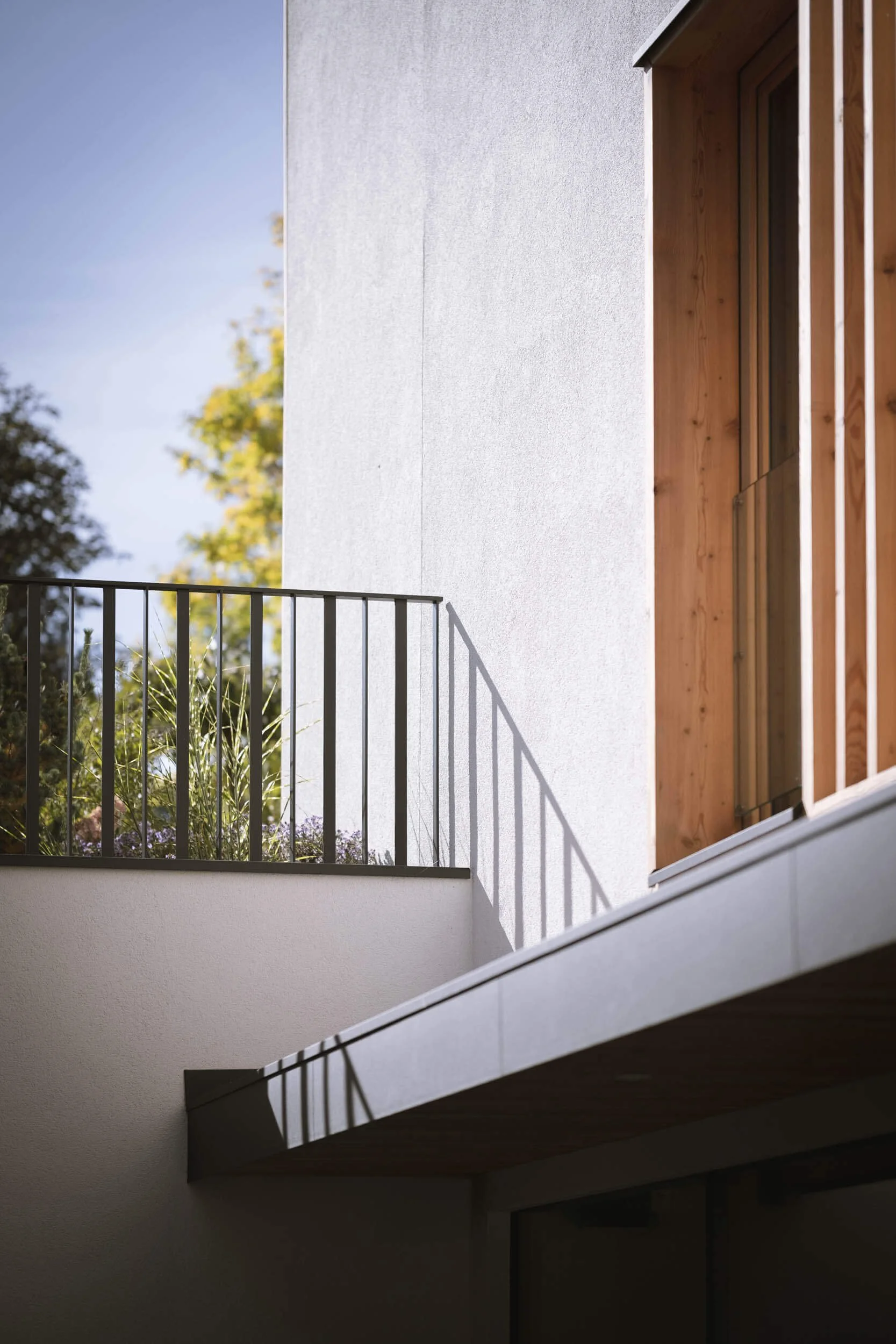 Balkon eines modernen Gebäudes mit weißen Wänden, schwarzer Geländer und Holzverkleidung, Schatten fallen durch die Geländerstäbe. Im Hintergrund sind Bäume sichtbar und der Himmel ist blassblau.