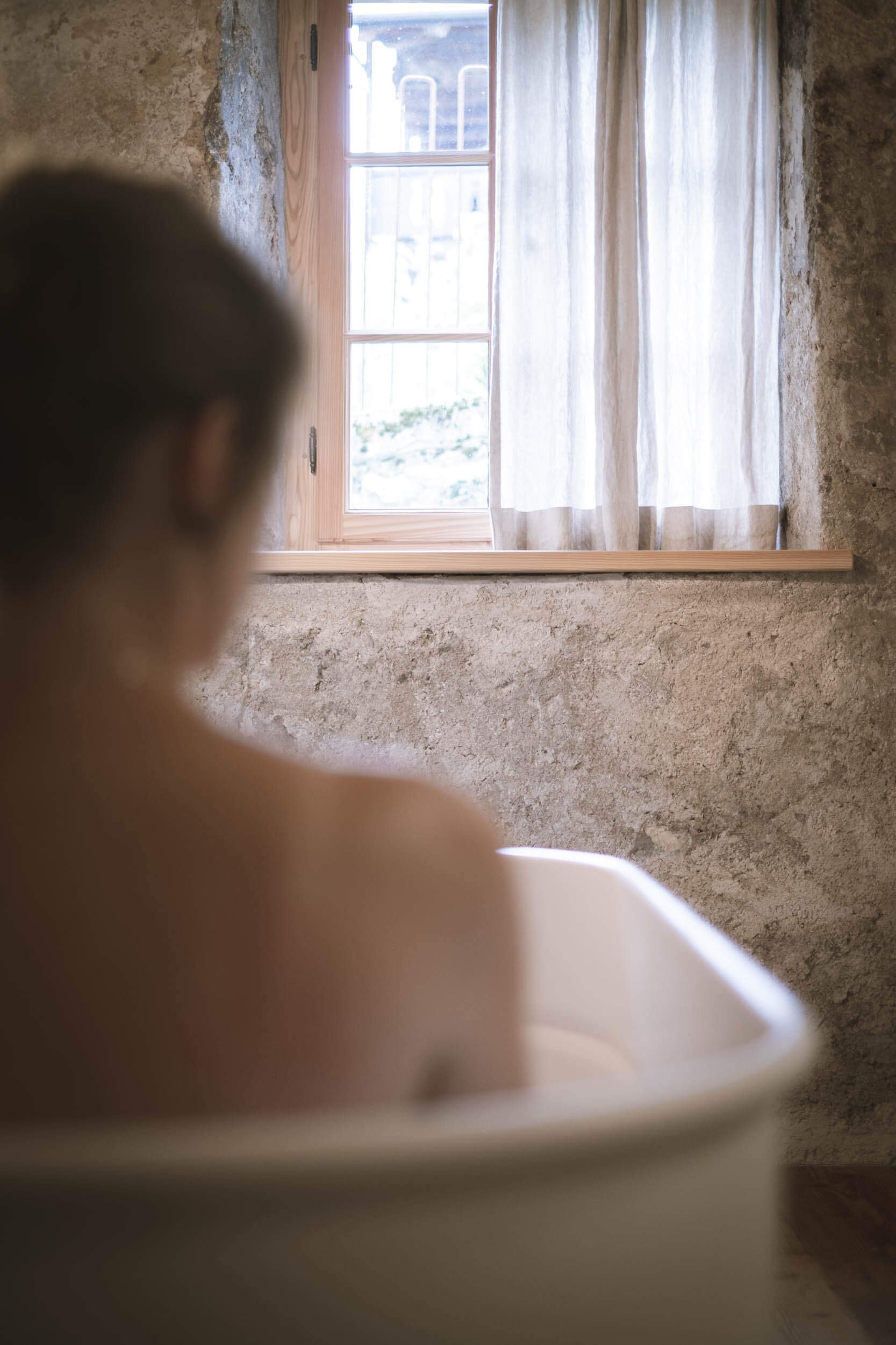 Eine Person sitzt in einer Badewanne vor einem Fenster mit weißen Vorhängen in einem Raum mit einer Steinwand.