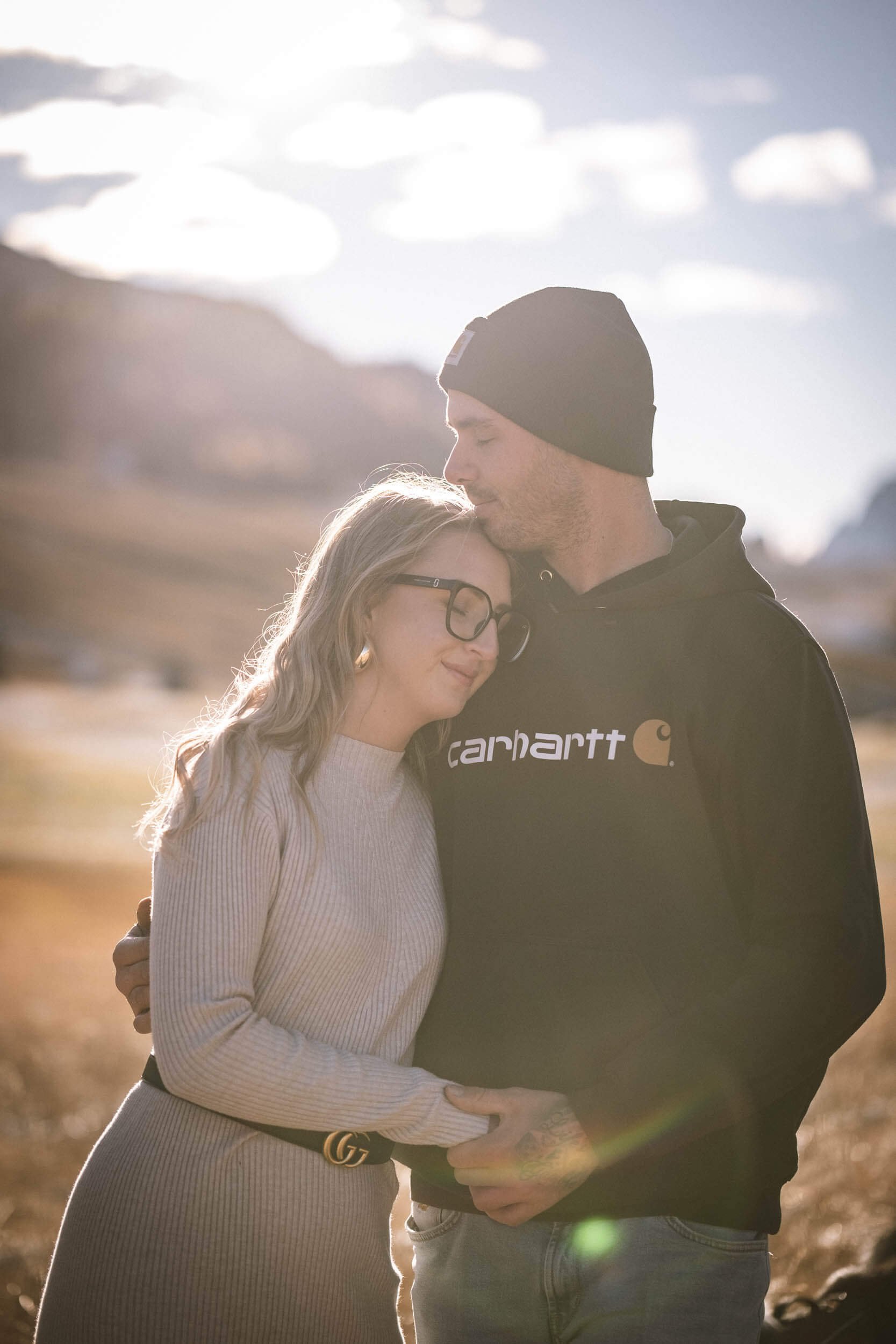 Ein Paar umarmt sich in einer lichten, sonnigen Landschaft bei Sonnenuntergang, das Mädchen trägt eine gestrickte Kleidung und Brille, der Mann trägt eine schwarze Kapuzenjacke mit Markenlogo.