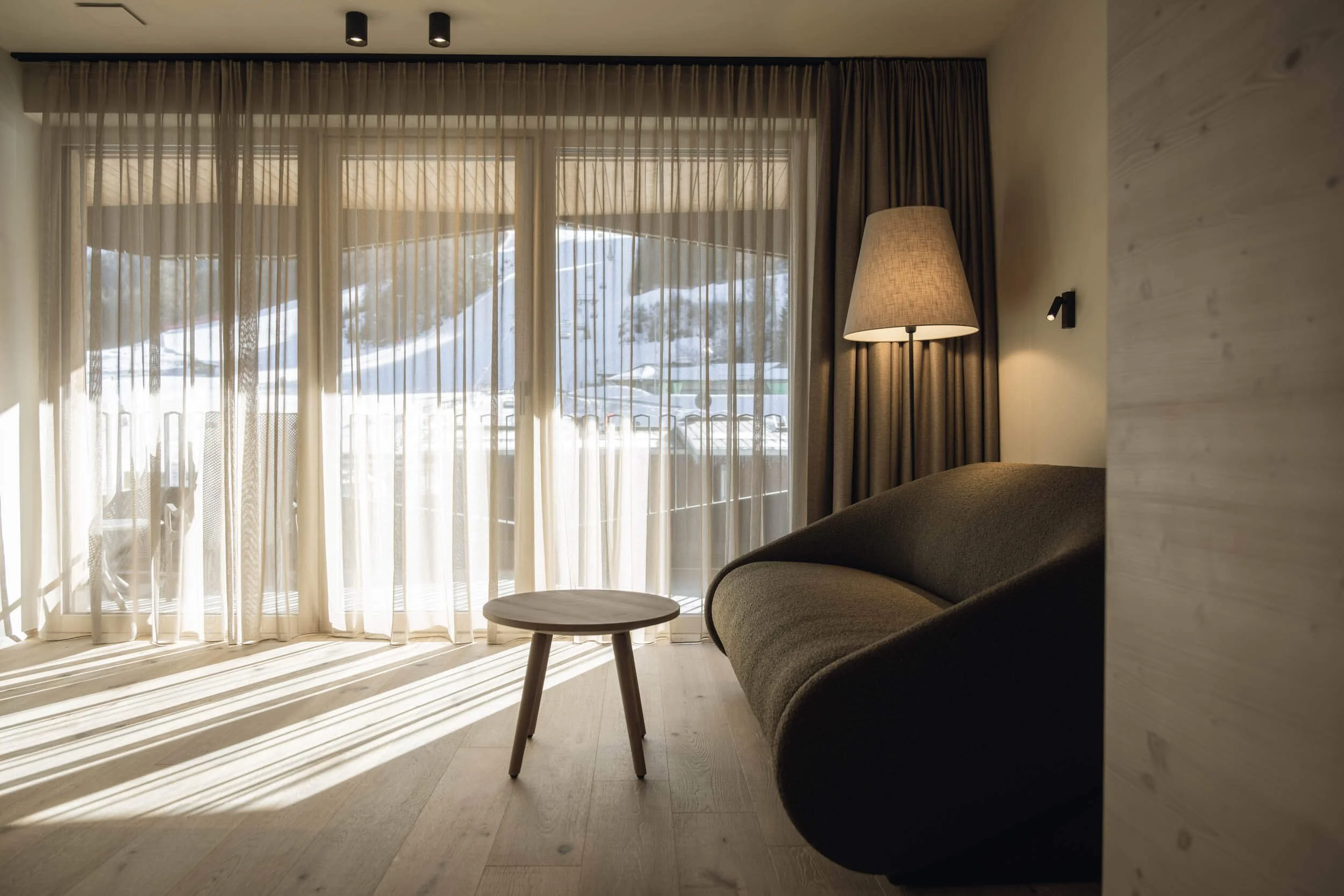 Gemütliches Wohnzimmer mit großen Fenster, durch das Schnee und Berge im Hintergrund sichtbar sind, ein Sofa, einen Stehlampe, einen kleinen runden Tisch und dicke Vorhänge.
