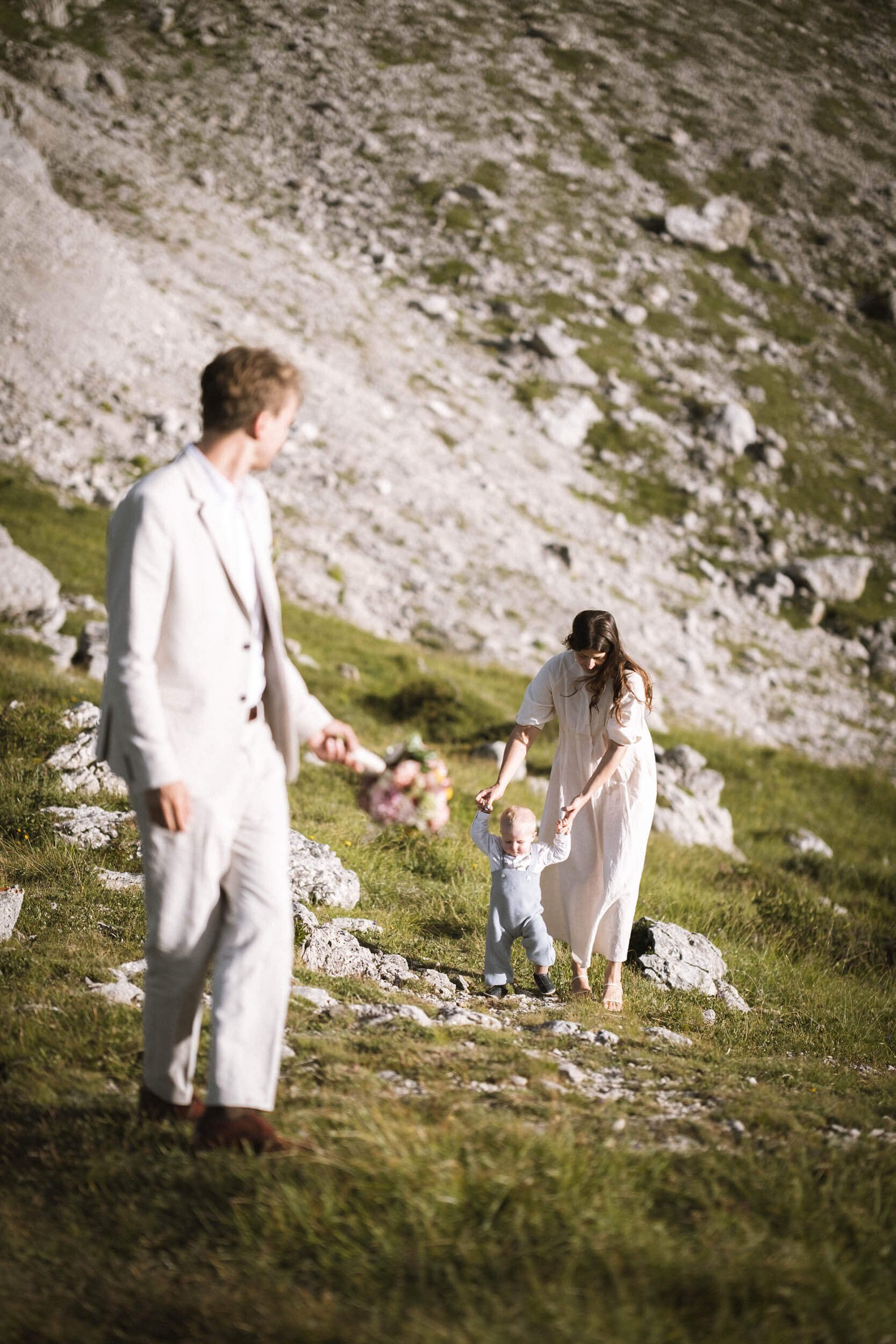 Familie beim Wandern in den Bergen, eine Frau hält ein Kind an den Händen, ein Mann steht im Vordergrund mit einem Blumenstrauß.