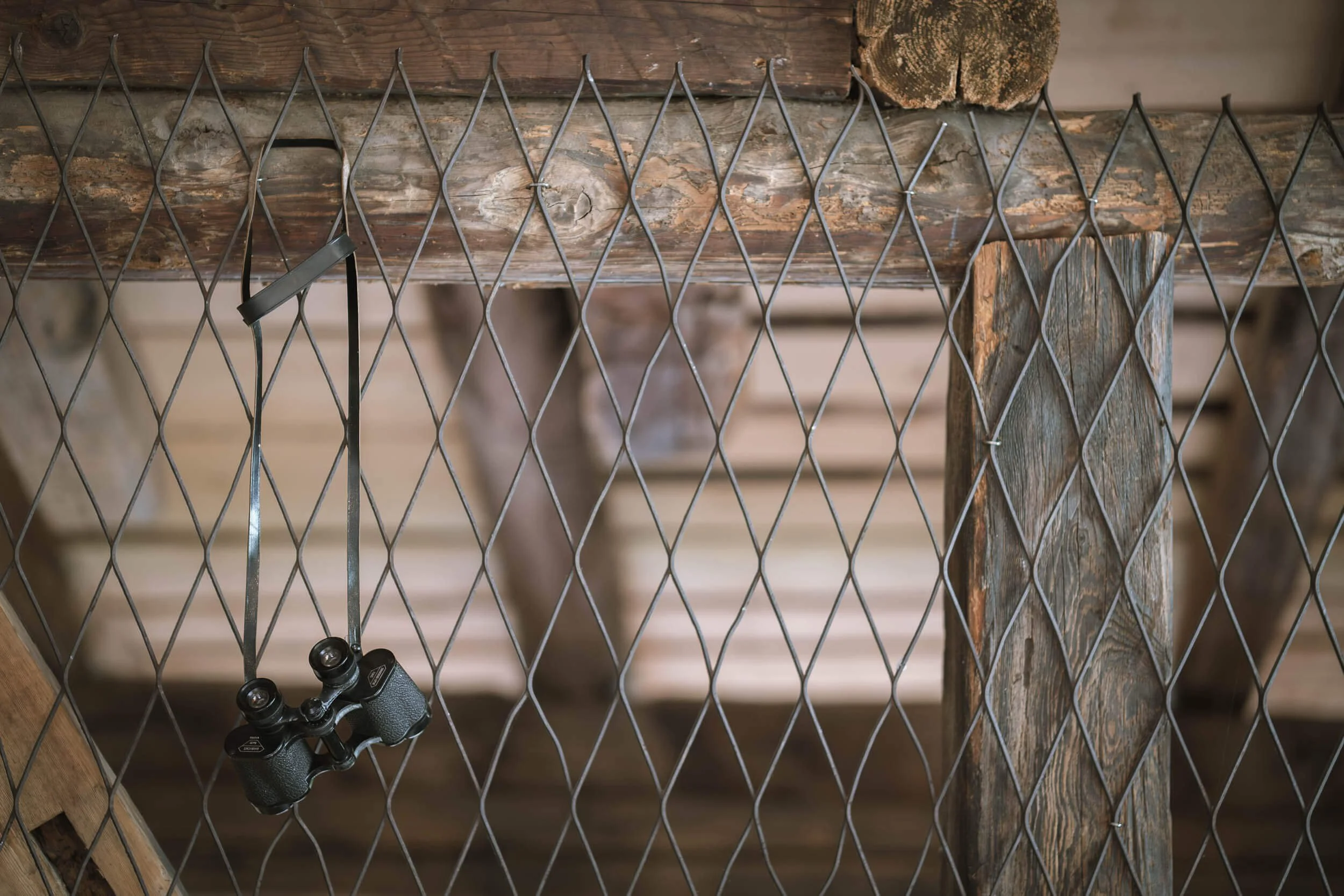 Ein Gitterzaun mit einem Fernglas daran, im Hintergrund Holzbohlen. Hinter dem Zaun sind Holzpfosten. Der Blick ist nach unten gerichtet.