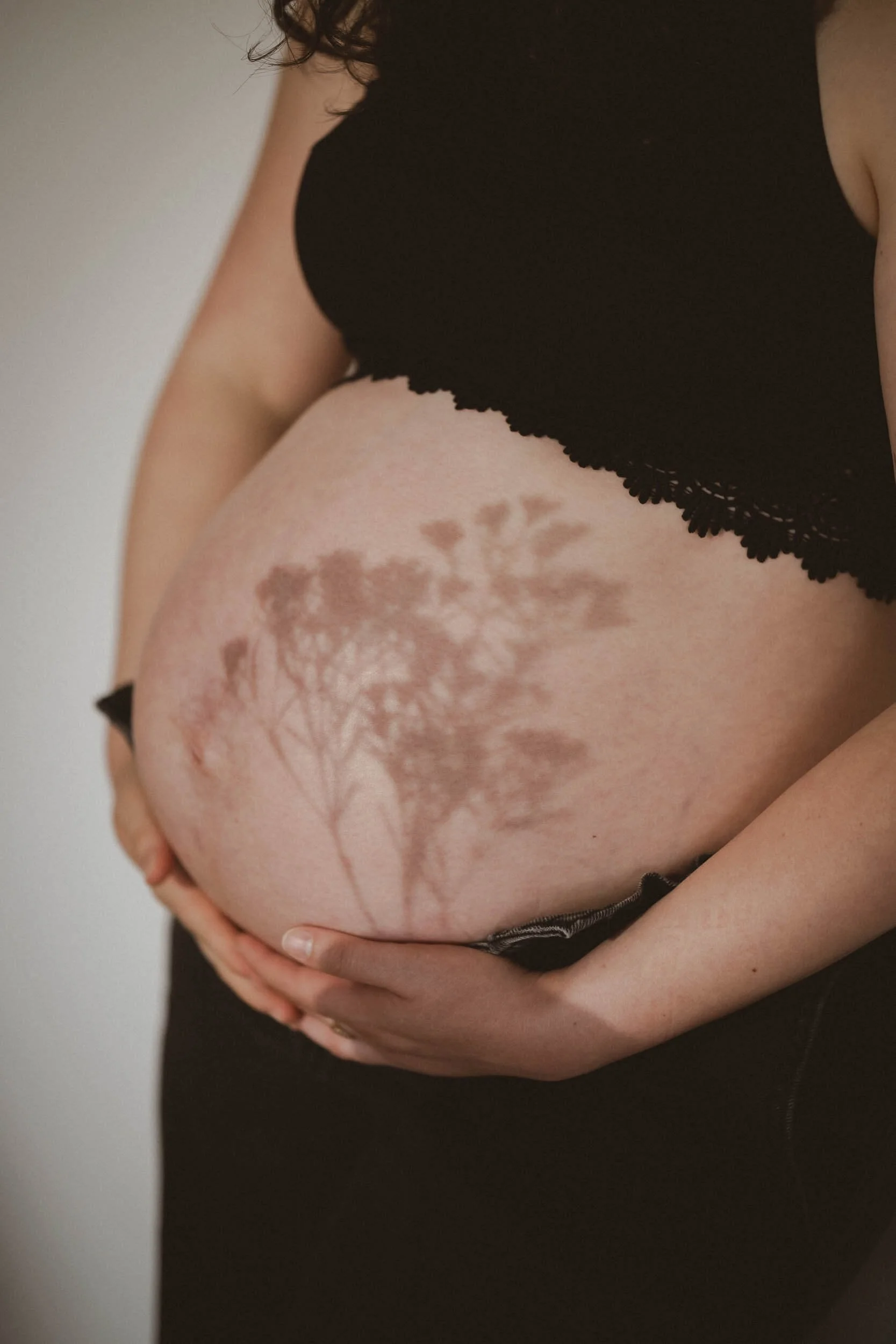 Schwangere Frau hält ihren Bauch, auf den der Schatten eines Baumes und Blätter fällt.