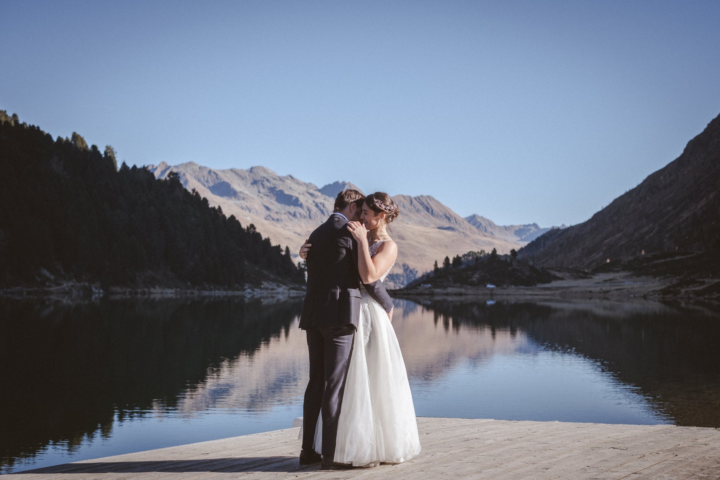 Ein Brautpaar umarmt sich vor einer Bergkulisse an einem See bei schönem Wetter.