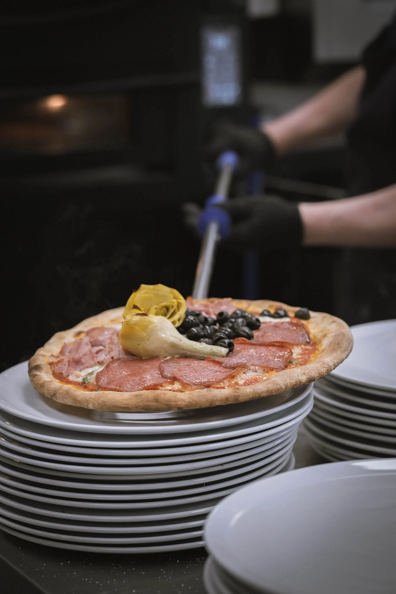 Eine Pizza mit schwarzen Oliven, Schinken und Salami, die auf einem weißen Teller auf einem Stapel von mehreren weißen Tellern liegt.