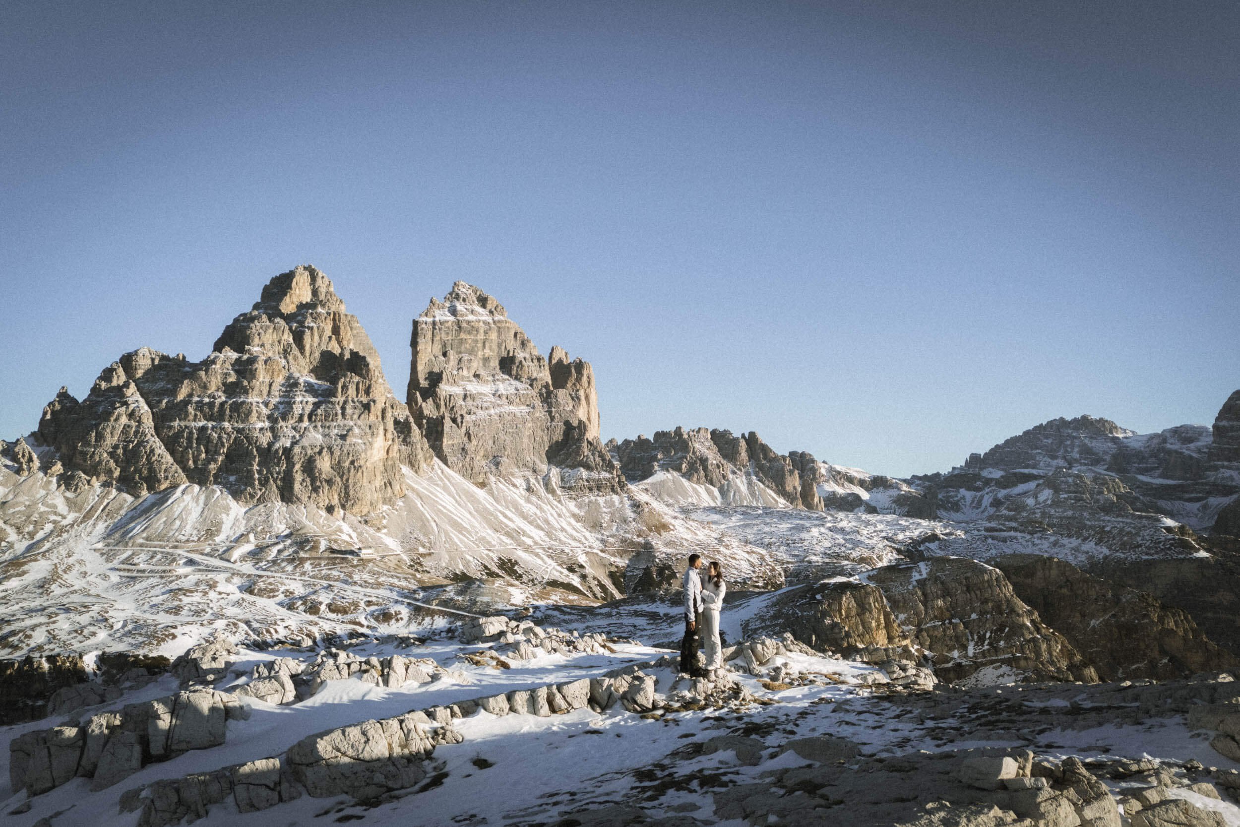Zwei Menschen in Winterkleidung stehen vor einer Bergkulisse mit schneebedeckten Gipfeln, sonnigem Himmel und felsigem Gelände.