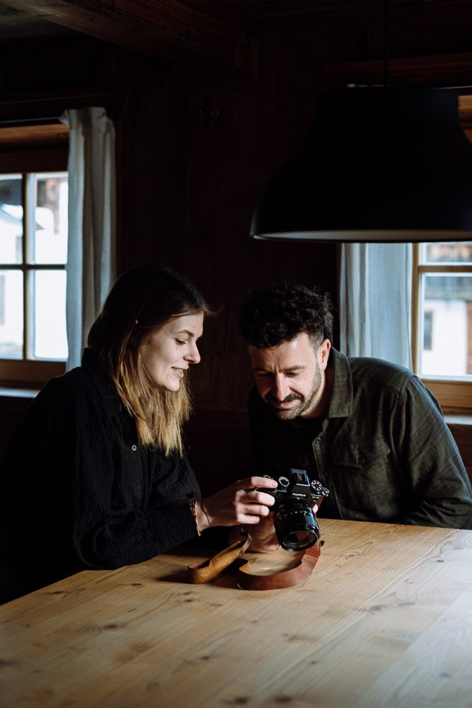 Zwei Menschen, eine Frau und ein Mann, betrachten eine Kamera auf einem Holztisch in einem Raum mit dunklen Wänden, großen Fenstern und weißen Vorhängen.