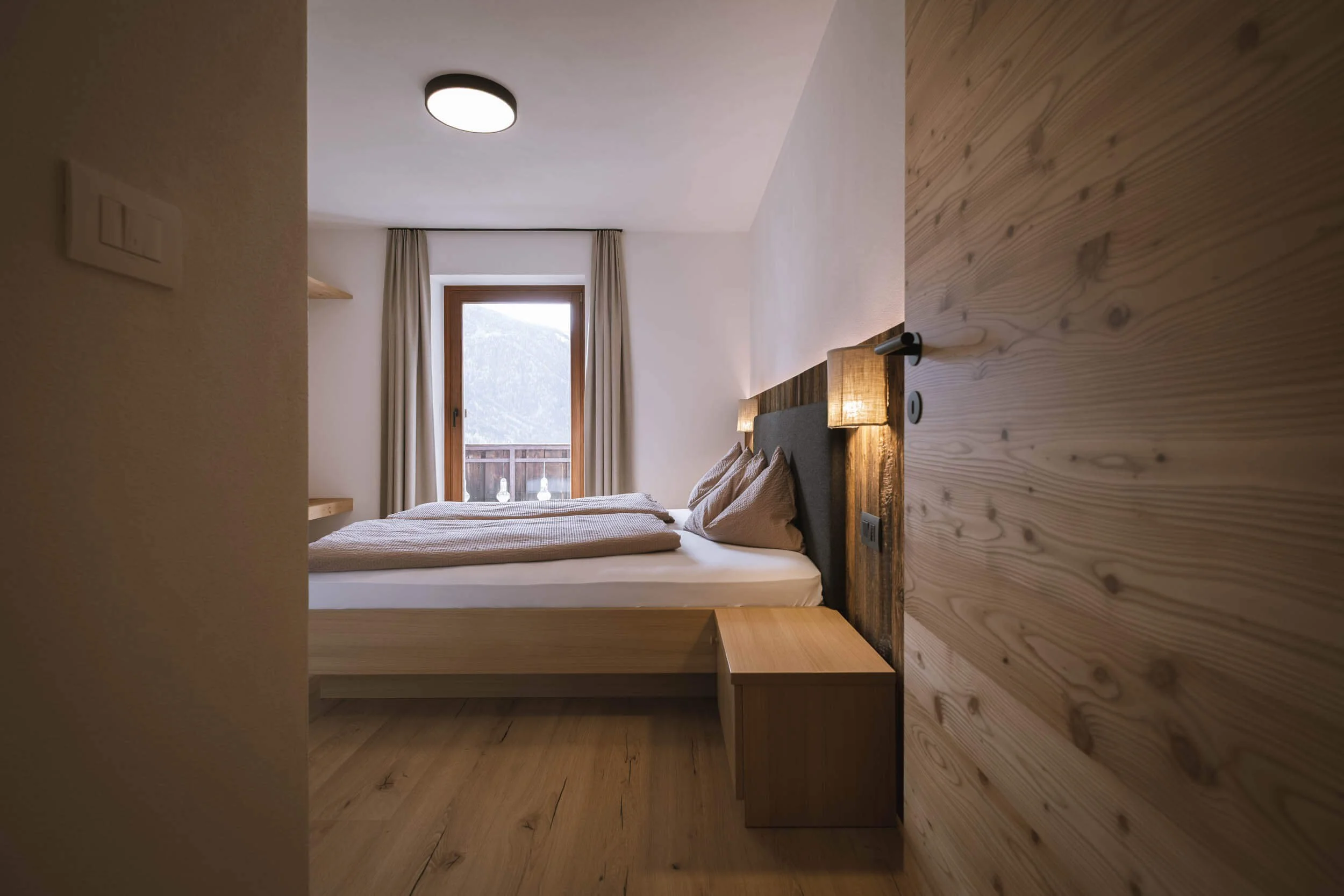 Ein Schlafzimmer mit einem Bett, Nachttisch und einer Fensterfront, die auf eine Berglandschaft blickt.