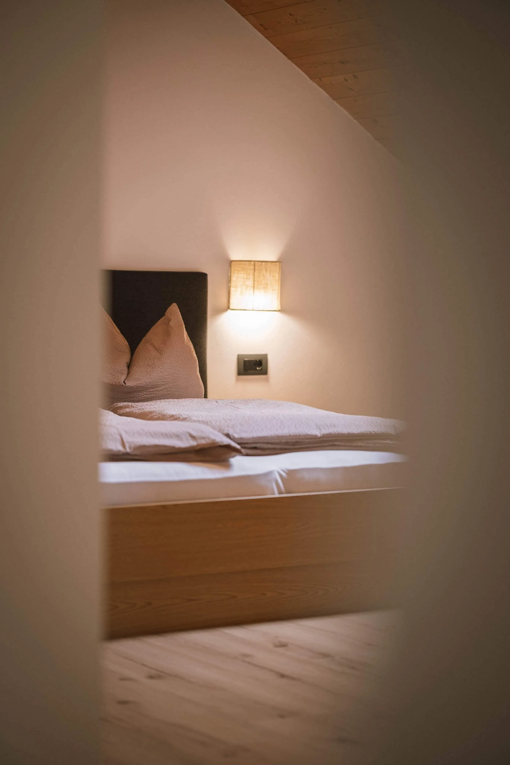 Ein Bett mit Kissen und Kopfteil, beleuchtet durch eine Wandlampe, gesehen durch ein offenes Türblatt in einem Schlafzimmer.