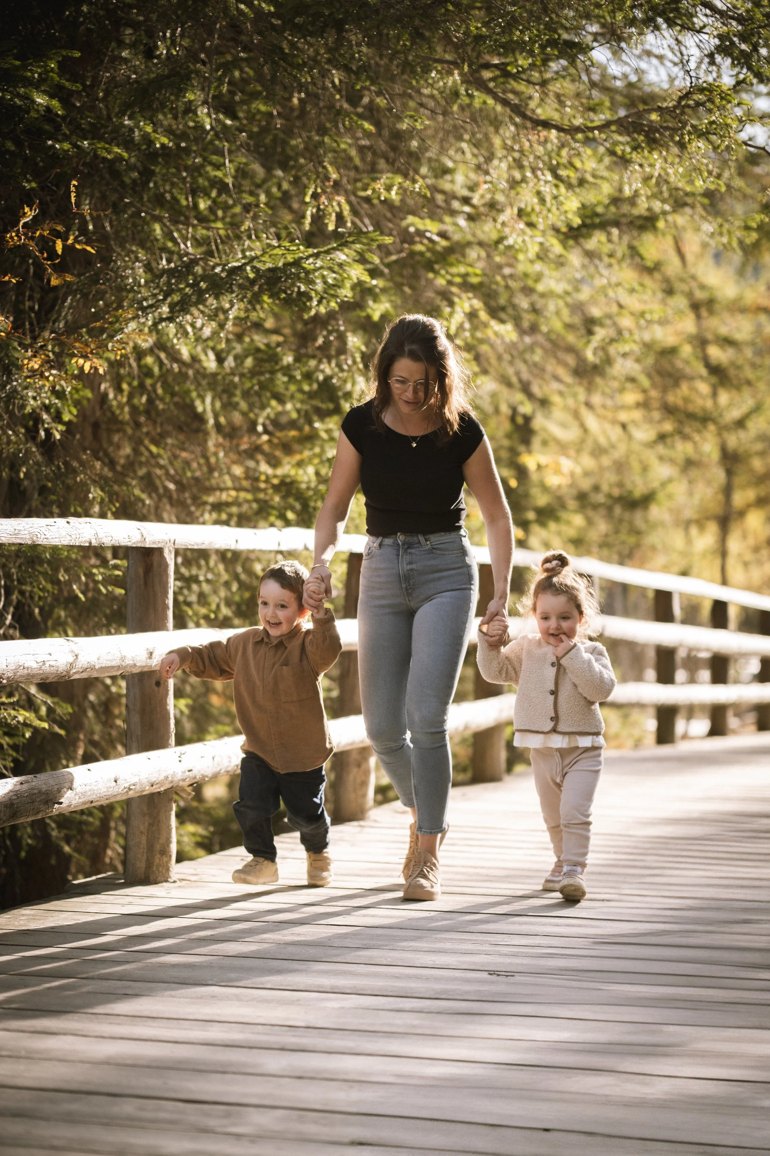 Eine Frau mit zwei kleinen Kindern, die auf einem hölzernen Weg in einem Baumpark spazieren gehen.