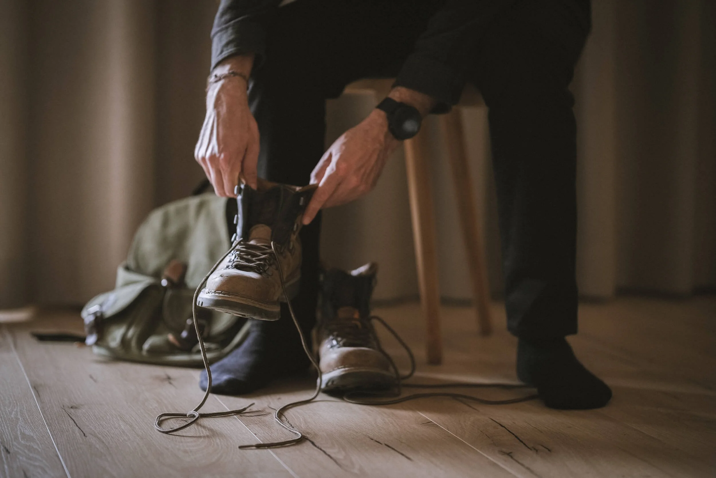 Person bindet Stiefel an, auf Holzfußboden, Rucksack im Hintergrund.