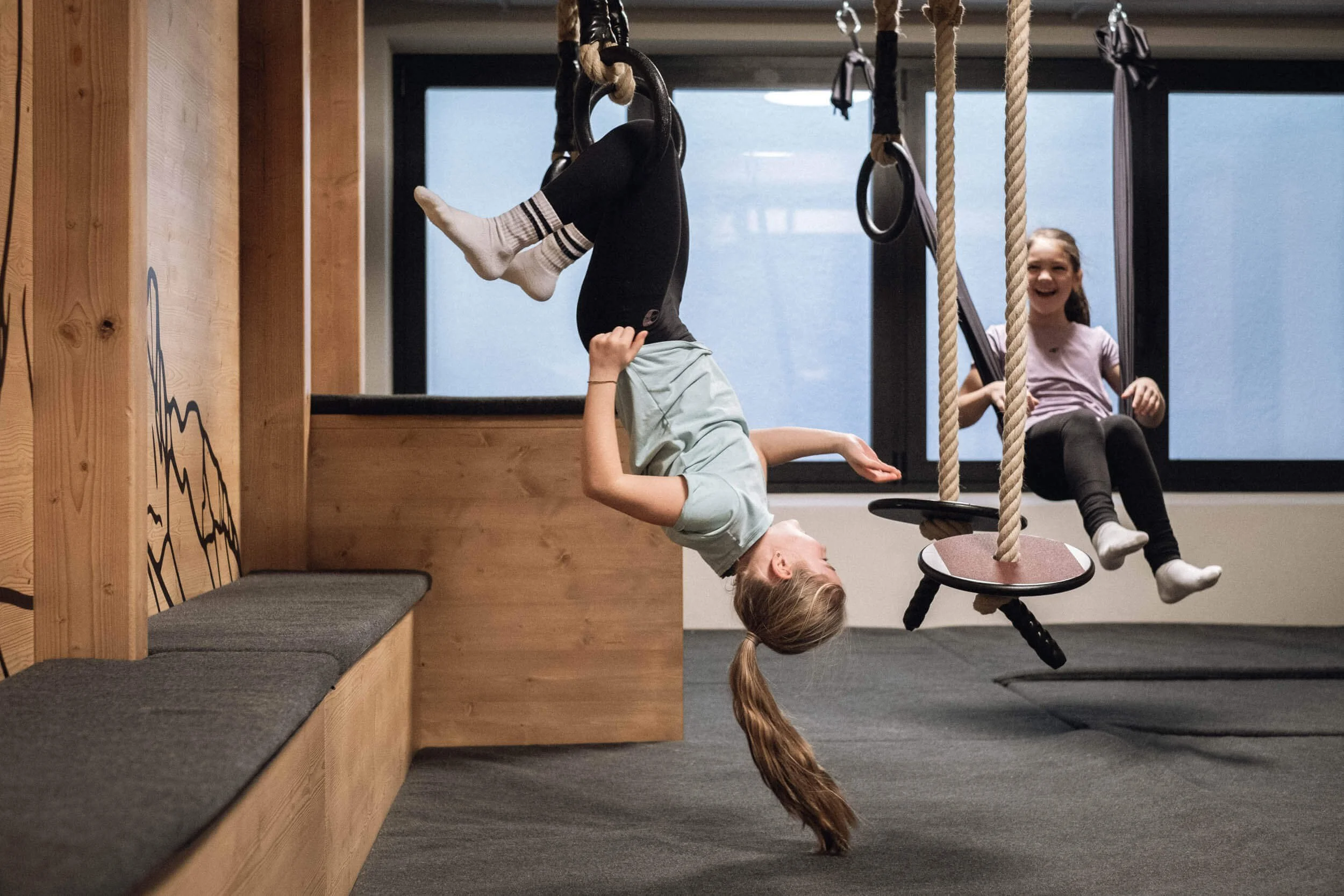 Zwei Kinder spielen in einem Indoor-Spielraum. Das Mädchen hängt kopfüber an mit Seilen befestigten Ringen, während das andere Mädchen auf einer Schaukel sitzt und lacht.