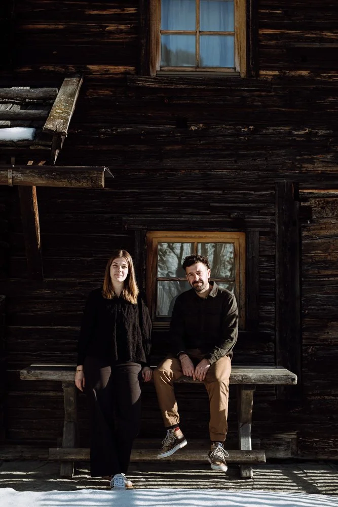 Ein Mann und eine Frau stehen vor einem alten, dunklen Holzhaus mit zwei Fenstern, im Winter mit Schnee. Der Mann sitzt auf einer Holzbank, die an der Hauswand angebracht ist, die Frau steht neben ihm.