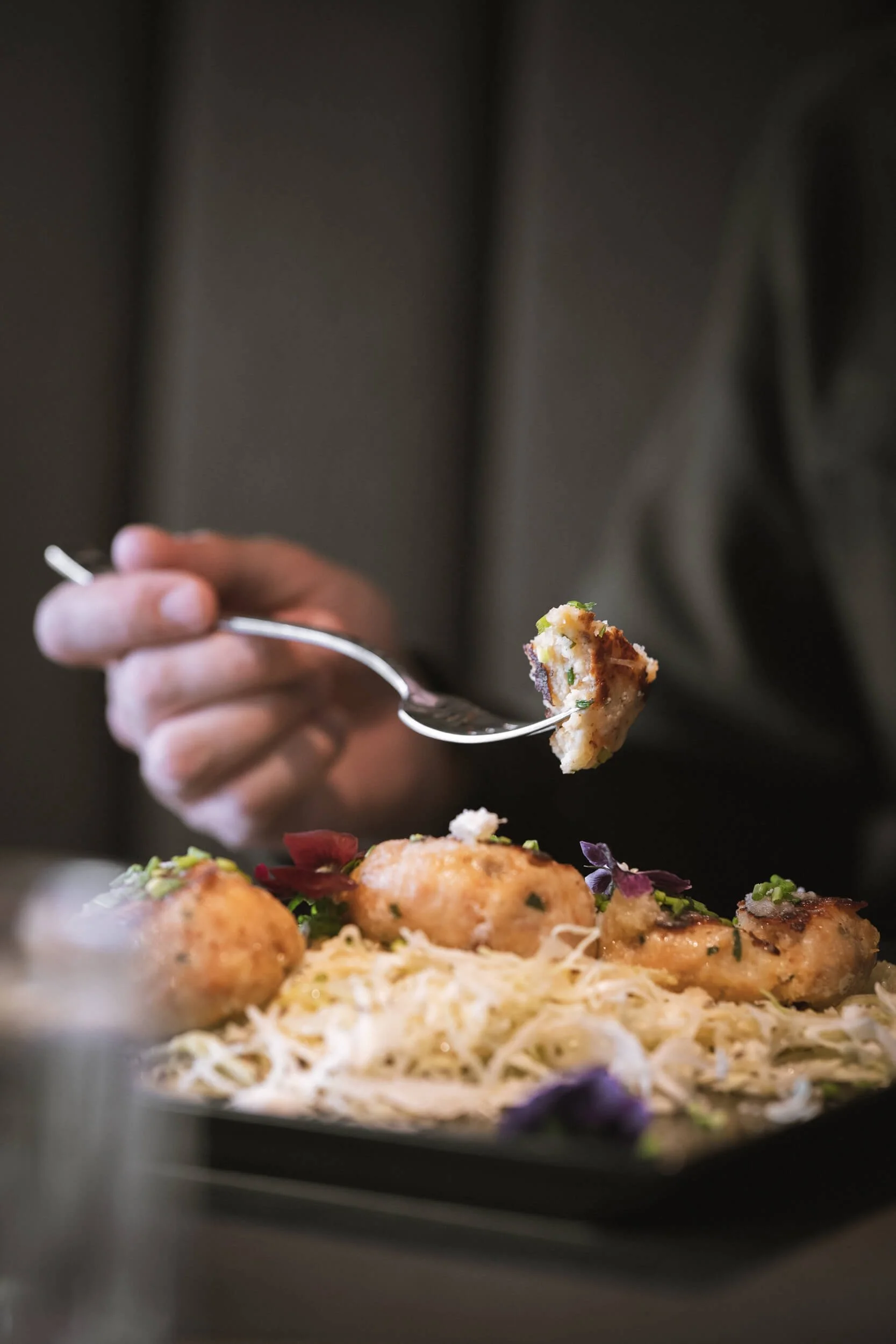 Nahaufnahme einer hand, die mit einer Gabel eine Portion Fisch mit Zitronen, Kräutern und Käse auf einem schwarzen Teller isst, im Hintergrund unscharf.