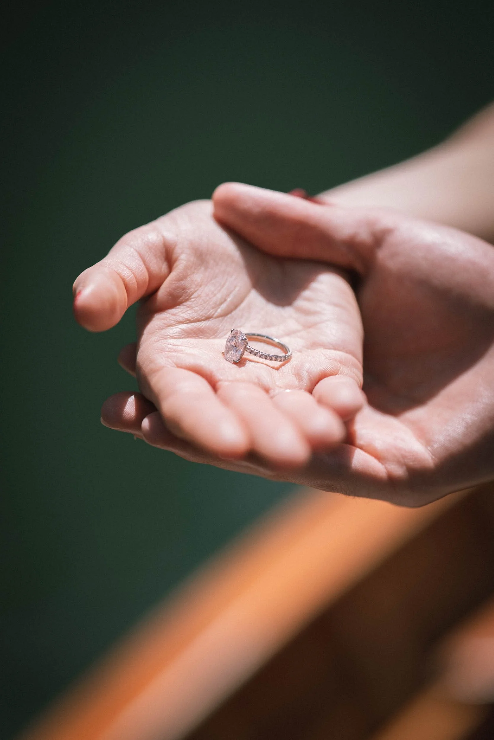 Eine Hand hält einen Verlobungsring mit großem Diamanten, das Licht fällt auf den Ring, im Hintergrund ist eine zweite Hand zu sehen.