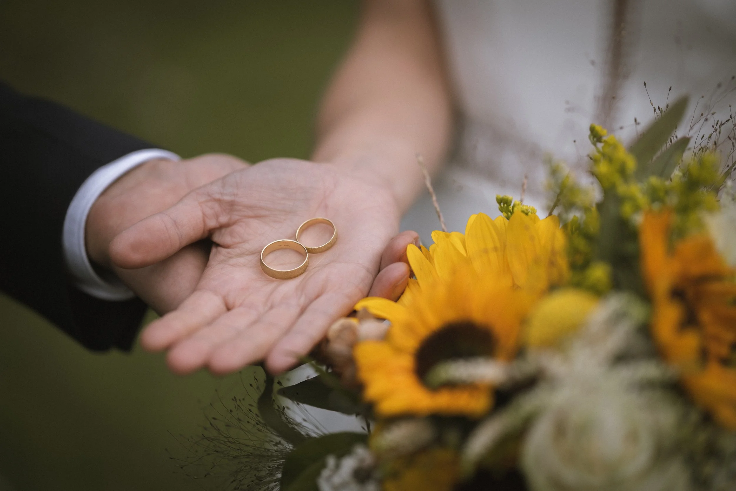 Hände eines Paars, das goldene Eheringe auf der offenen Hand hält, neben Sonnenblumen und anderen Blumen bei einer Zeremonie.
