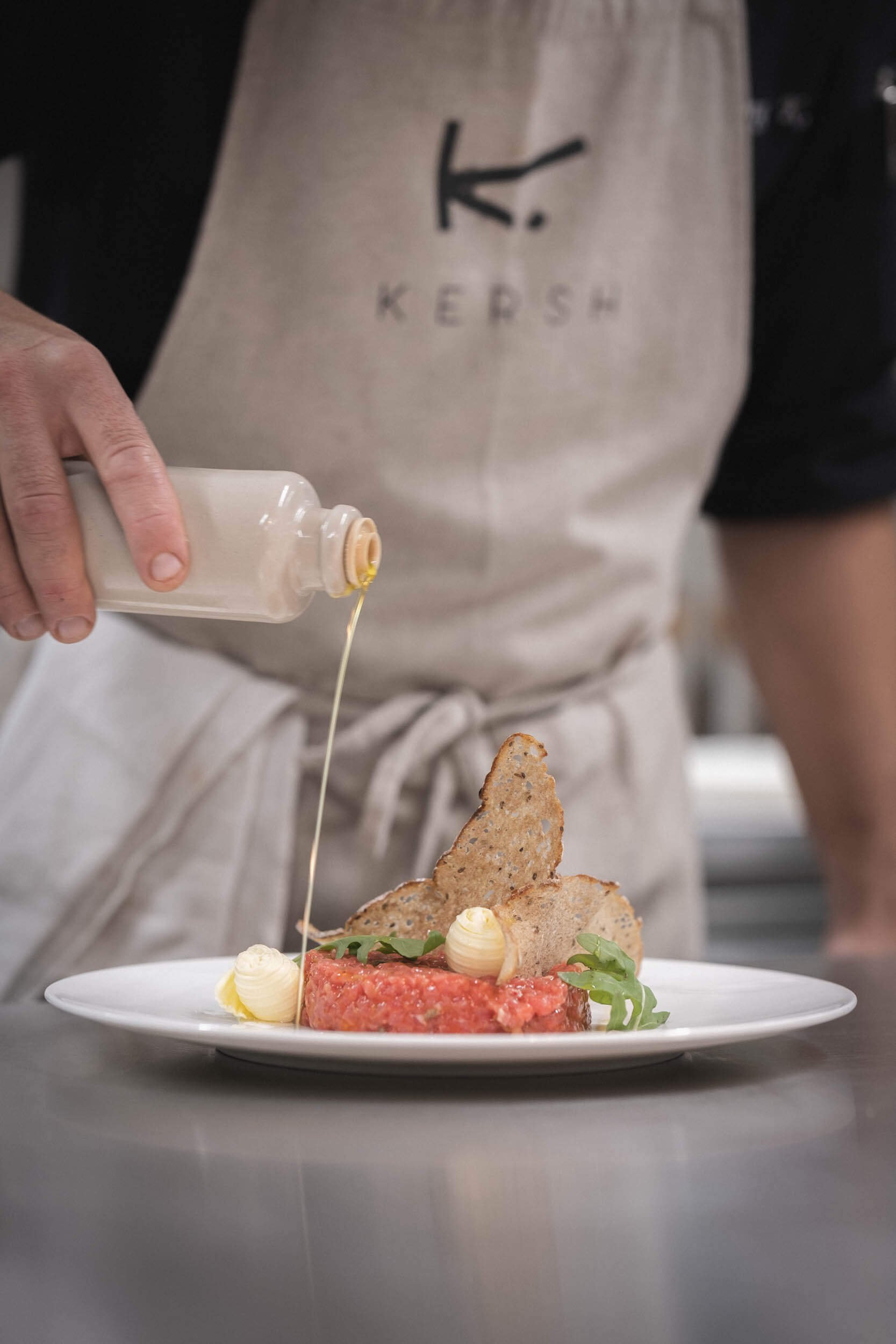 Ein Koch trägt eine Schürze mit dem Logo 'Kersh' und dekoriert ein Gericht mit Olivenöl.