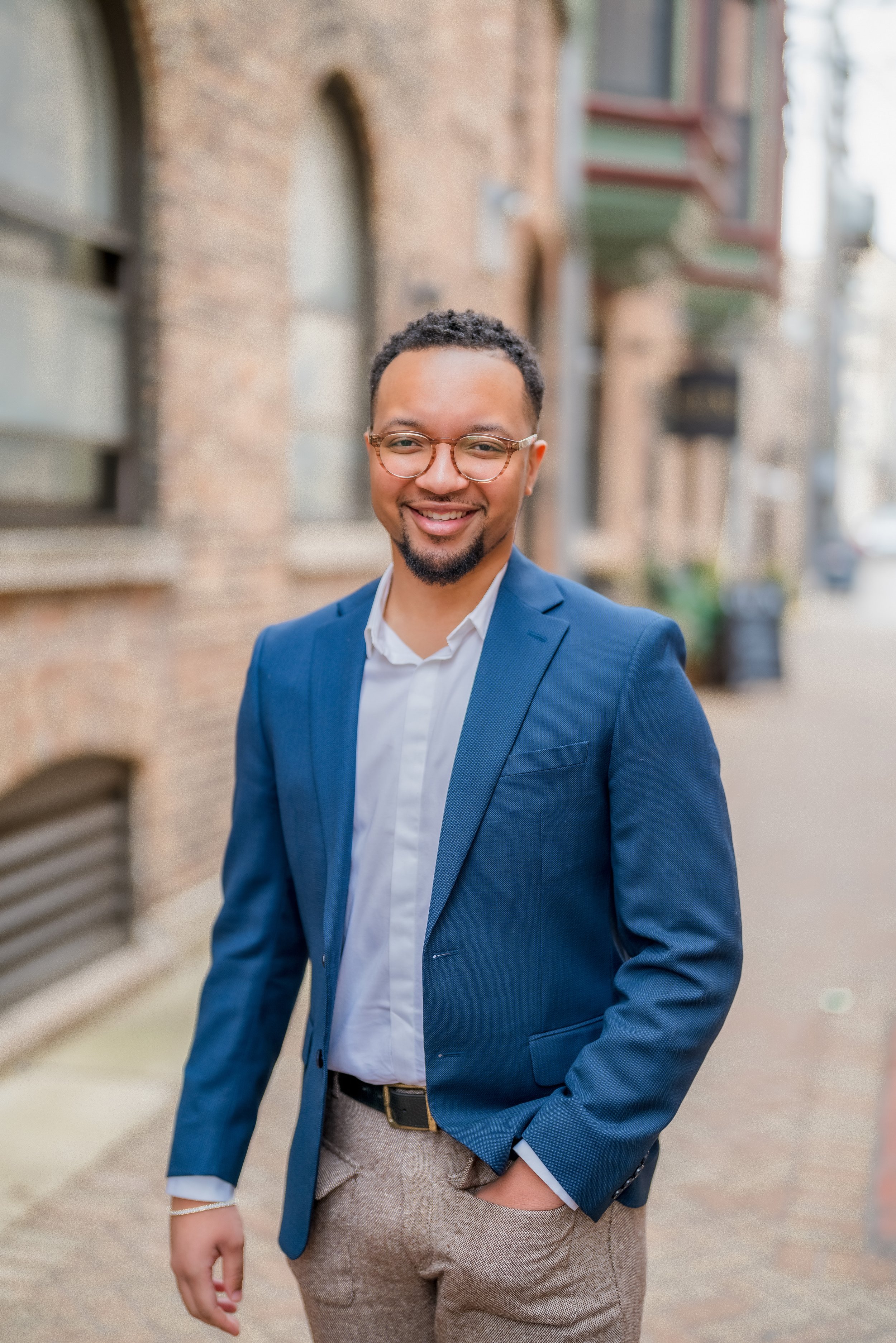 Smiling man in blue blazer and glasses standing on city sidewalk with brick building background.
