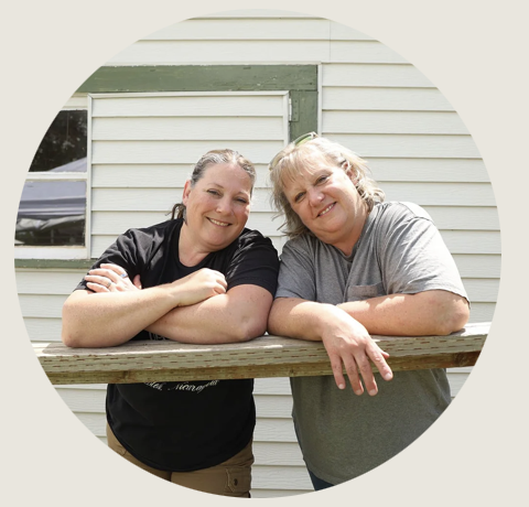 Two women lean comfortably on a wooden railing outside a light-colored building, smiling at the camera. Dressed in t-shirts and jeans, they appear relaxed and friendly.