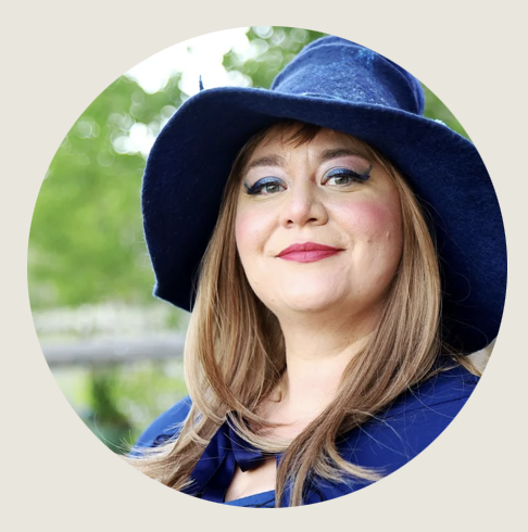 Casper-A woman in a deep blue outfit and wide-brimmed hat smiles softly at the camera. She wears bold lipstick and a confident gaze. A blurred green background suggests she’s standing in a natural setting.