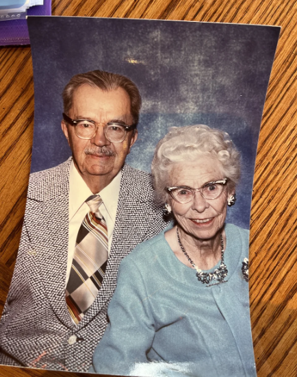 Cheyenne- An older couple poses together for a formal portrait. The man wears glasses, a patterned blazer, and striped tie; the woman, also in glasses, wears a light blue sweater with matching necklace. They sit closely, smiling.