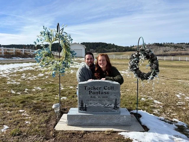 A man and woman sit behind a gravestone that reads "Tucker Colt Pantuso," with two wreaths on stands nearby, in a grassy cemetery with snow patches and a rural landscape.