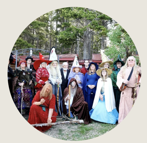 Casper- A large group of people dressed as wizards, witches, and mystical characters gather outdoors in front of a rustic cabin surrounded by pine trees.