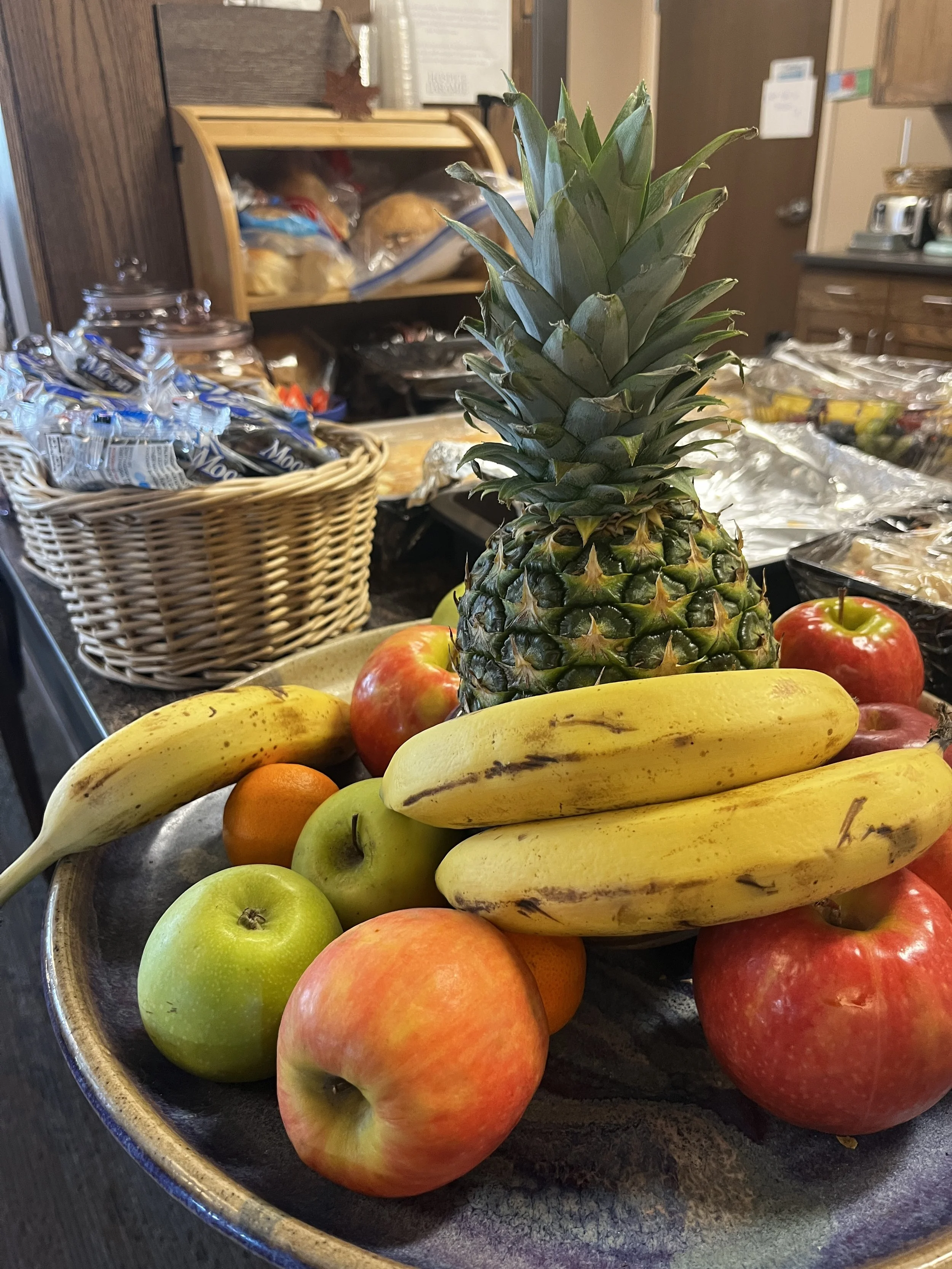 A large bowl filled with a fresh pineapple, yellow bananas, red apples, green apples, and small oranges, set on a counter with a wicker basket. of snacks in the background