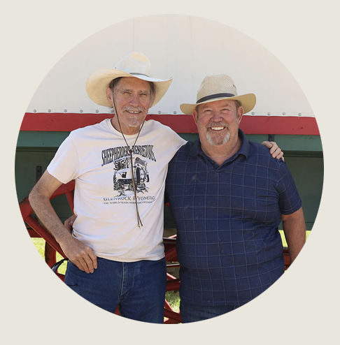 Two smiling men in hats stand arm-in-arm outdoors. One wears a white cowboy hat and “Sheepherders Rendezvous” T-shirt; the other wears a straw hat and blue plaid shirt. They stand in front of a green and red sheep wagon with a white cover.