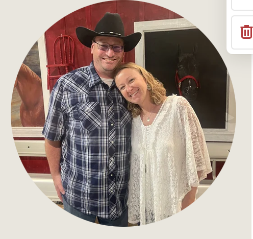 Cheyenne- A smiling couple stands close together indoors, the woman resting her head on the man’s shoulder. He wears a cowboy hat and blue plaid shirt; she wears a white lace blouse. Behind them is a small red chair and framed horse portrait.